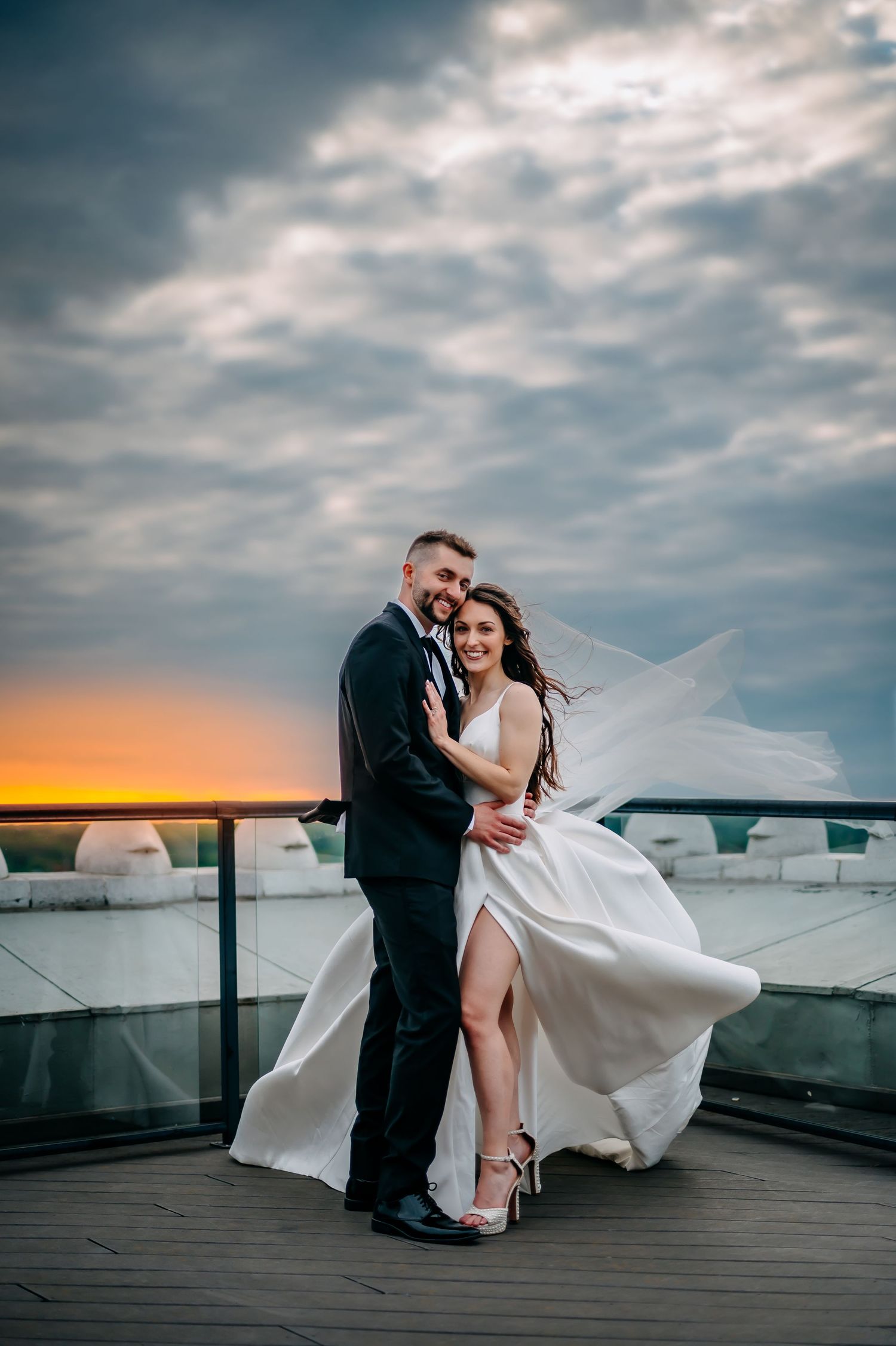 Couple in wedding attire embrace on rooftop terrace with dramatic sunset sky and flowing white dress Grand Rapids Michigan Mckay
