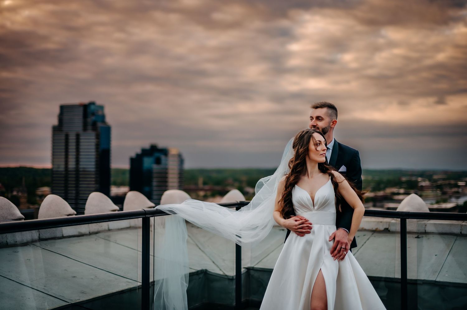 A romantic couple in wedding attire embrace on a rooftop terrace at sunset with city skyline in the background Ballroom At Mckay Tower