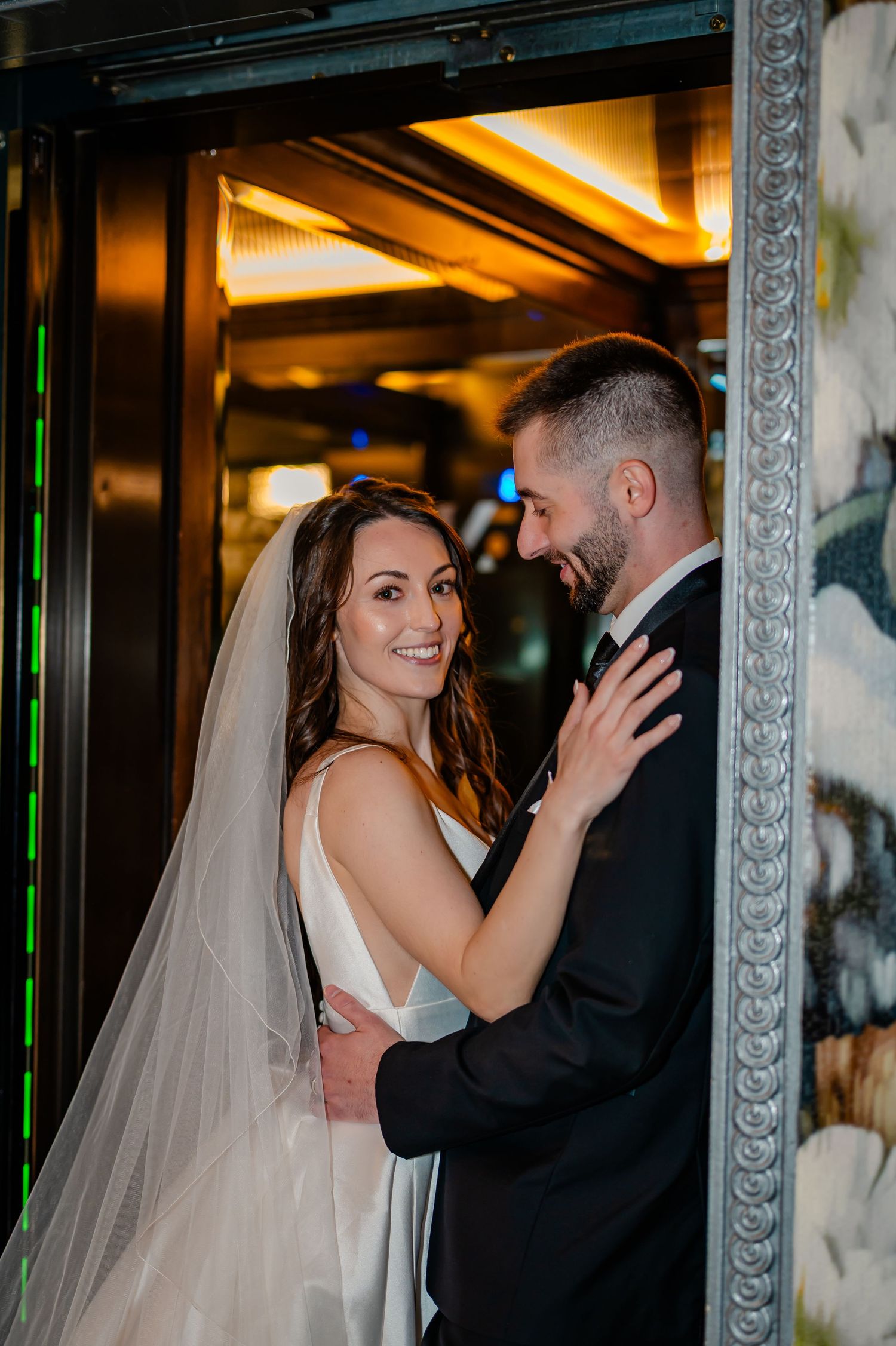 Bride in white gown and veil embraces groom in black suit, smiling in elegant venue doorway.