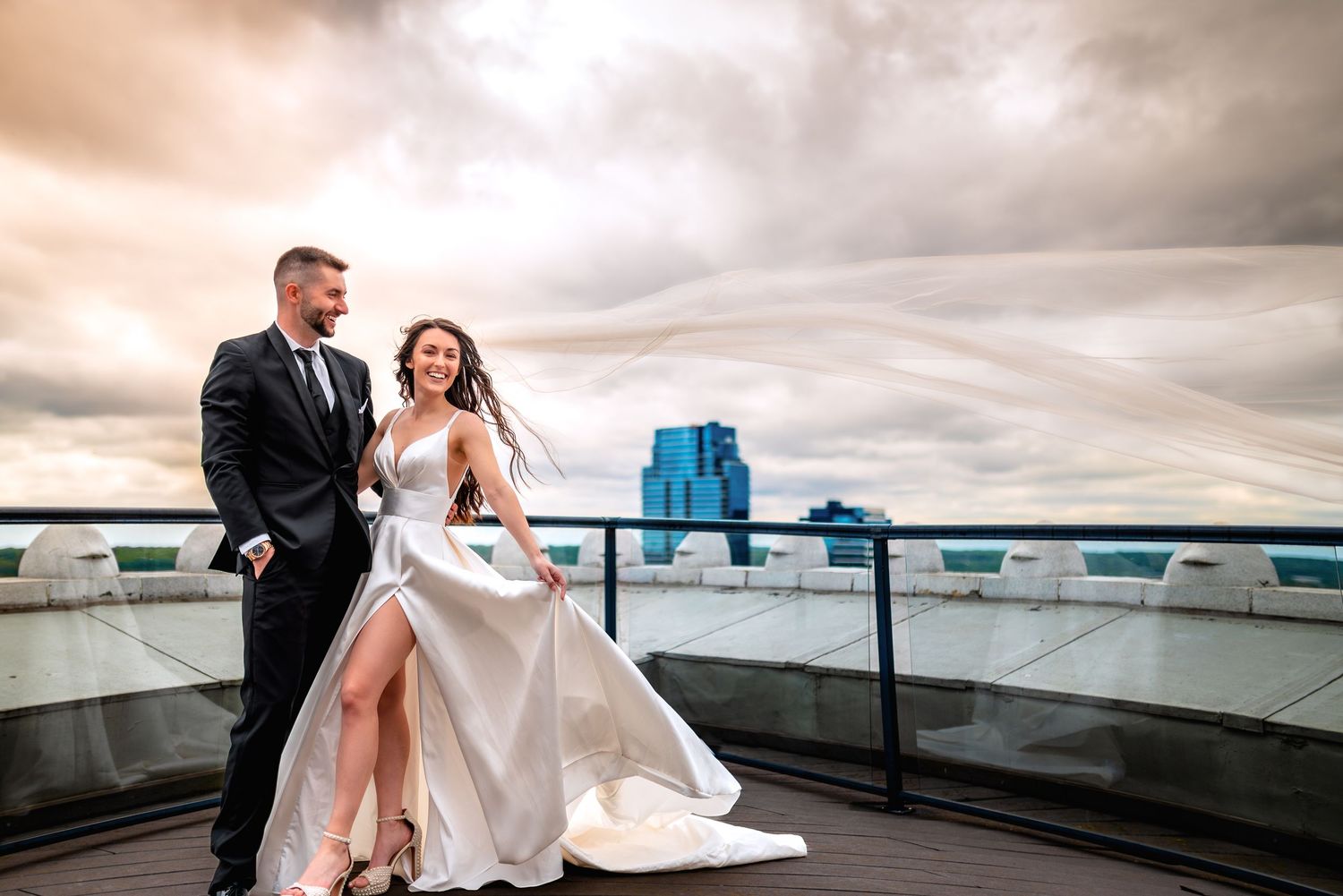 Bride and groom walk on rooftop terrace under dramatic cloudy sky with city skyline backdrop.