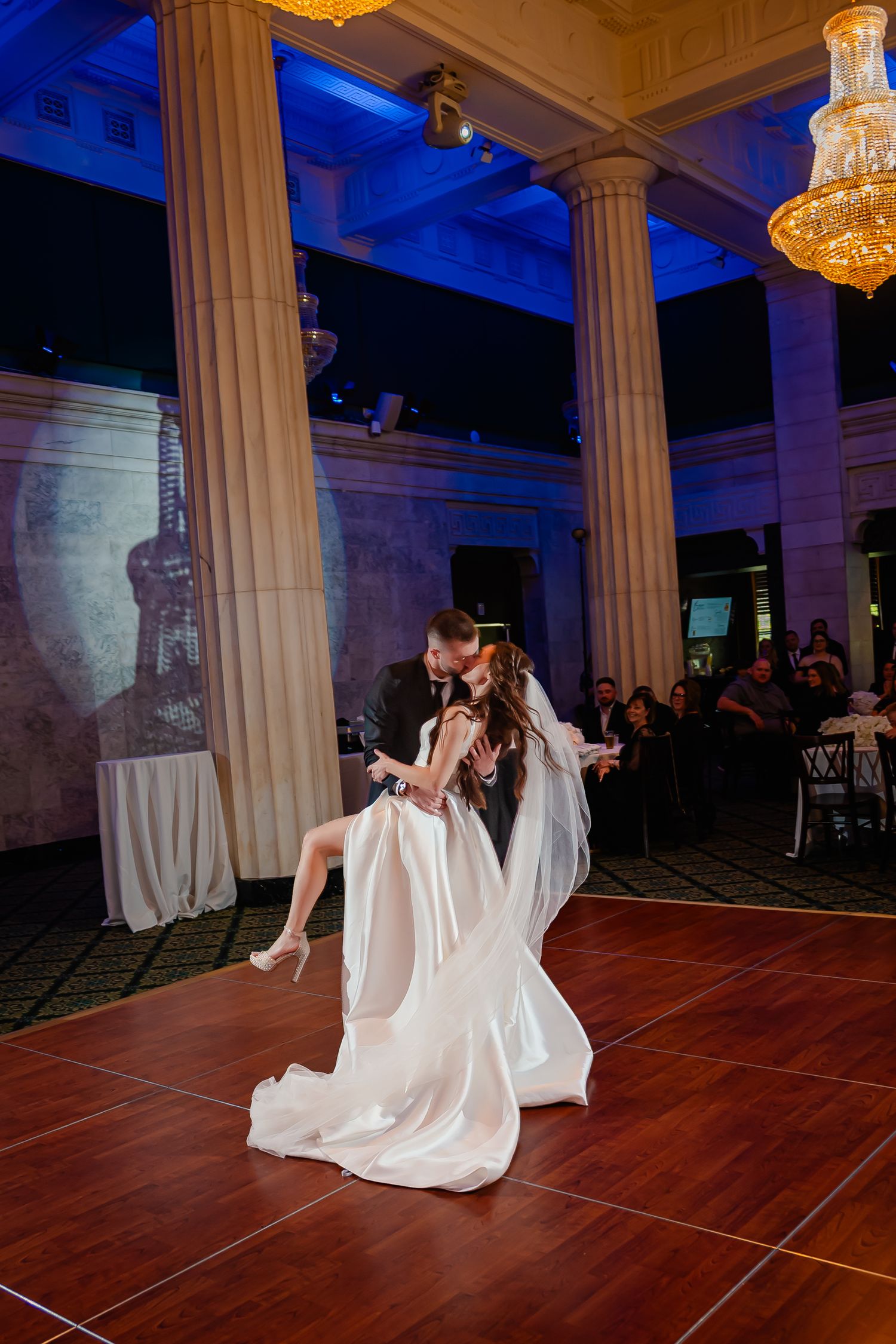 Bride and groom share romantic first dance in elegant ballroom with crystal chandeliers and blue uplighting.