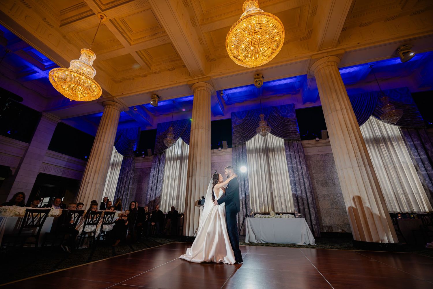 Bride and groom share first dance in elegant ballroom with grand columns and blue uplighting at wedding reception.