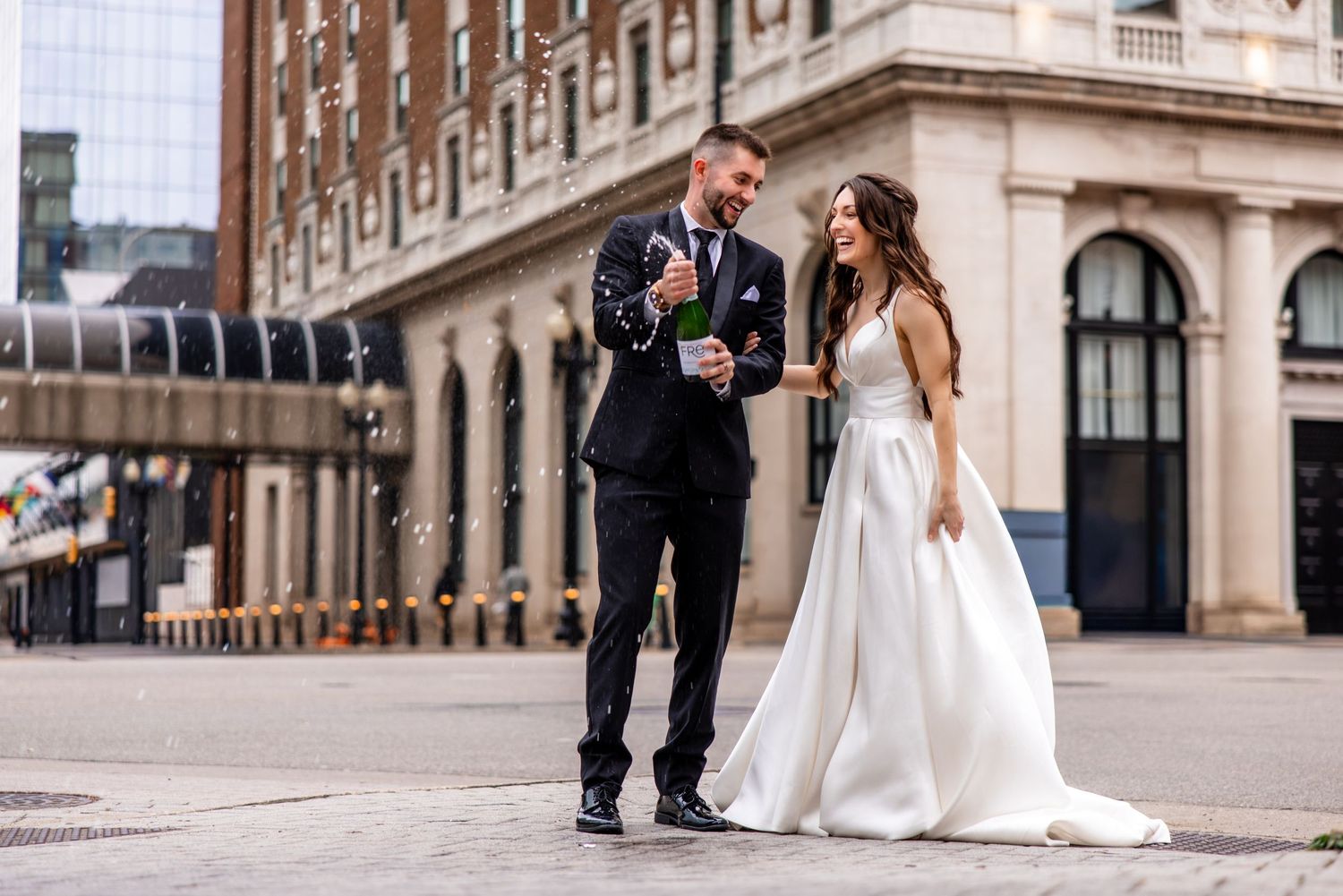 A bride and groom laugh together outdoors, popping champagne in front of a classic brick building on their wedding day.