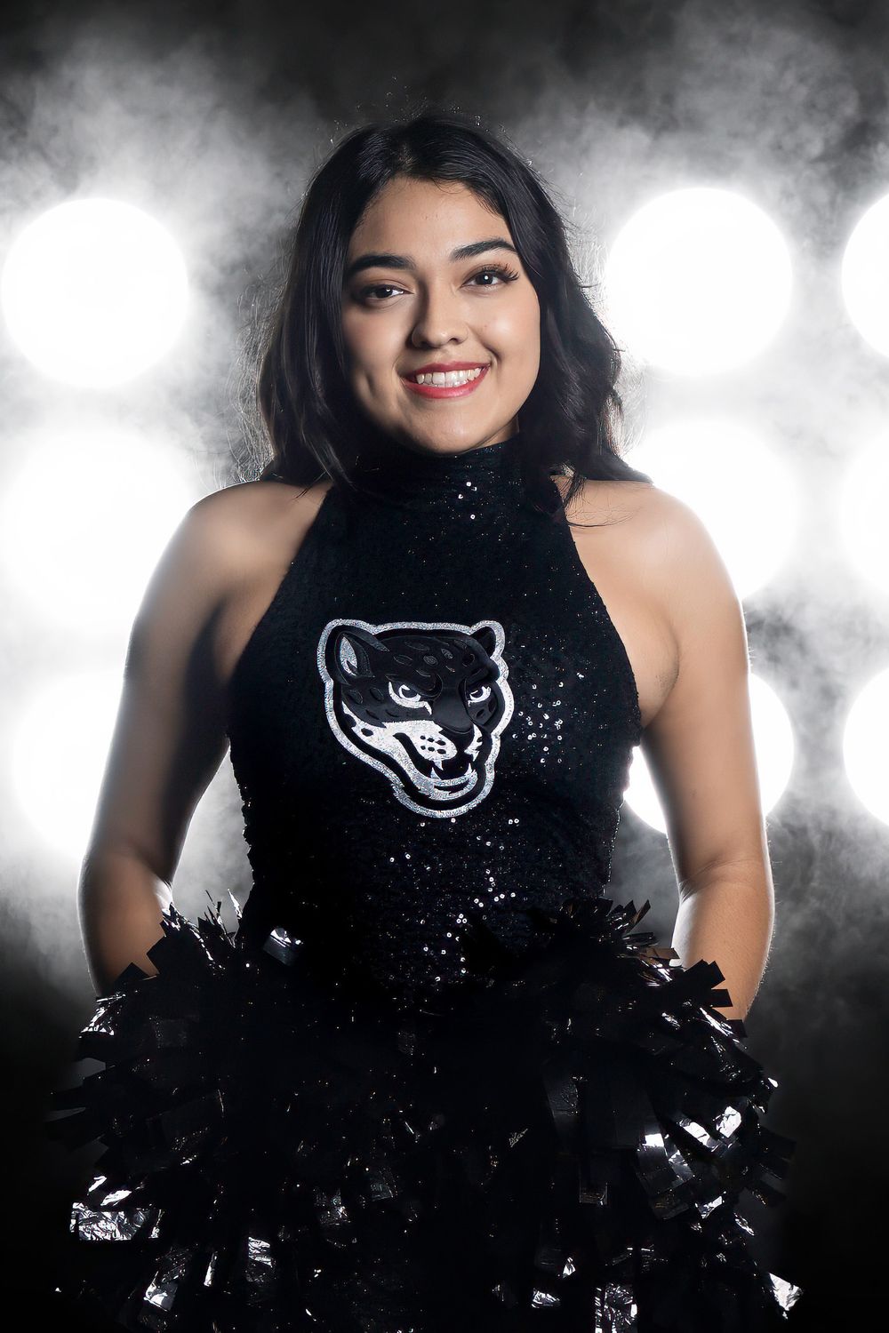 A cheerleader poses in a sparkly black halter uniform featuring a panther mascot logo against a smoky backdrop.