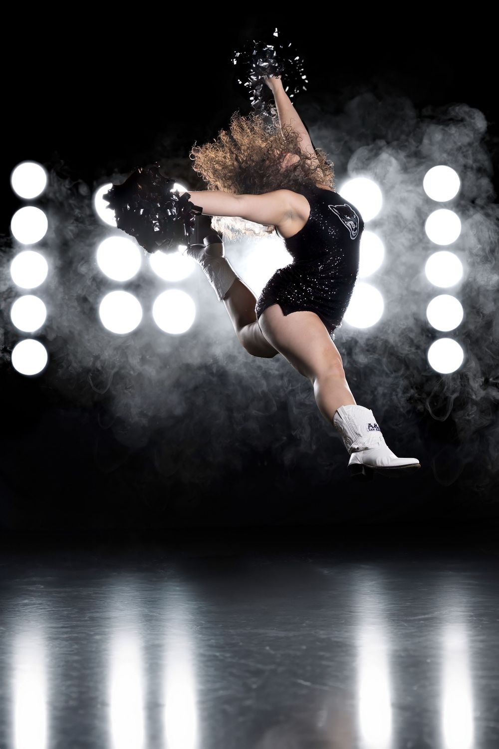 Dynamic figure skater in black leotard performs graceful leap against dramatic backlit stage with bokeh lights and smoke effects.