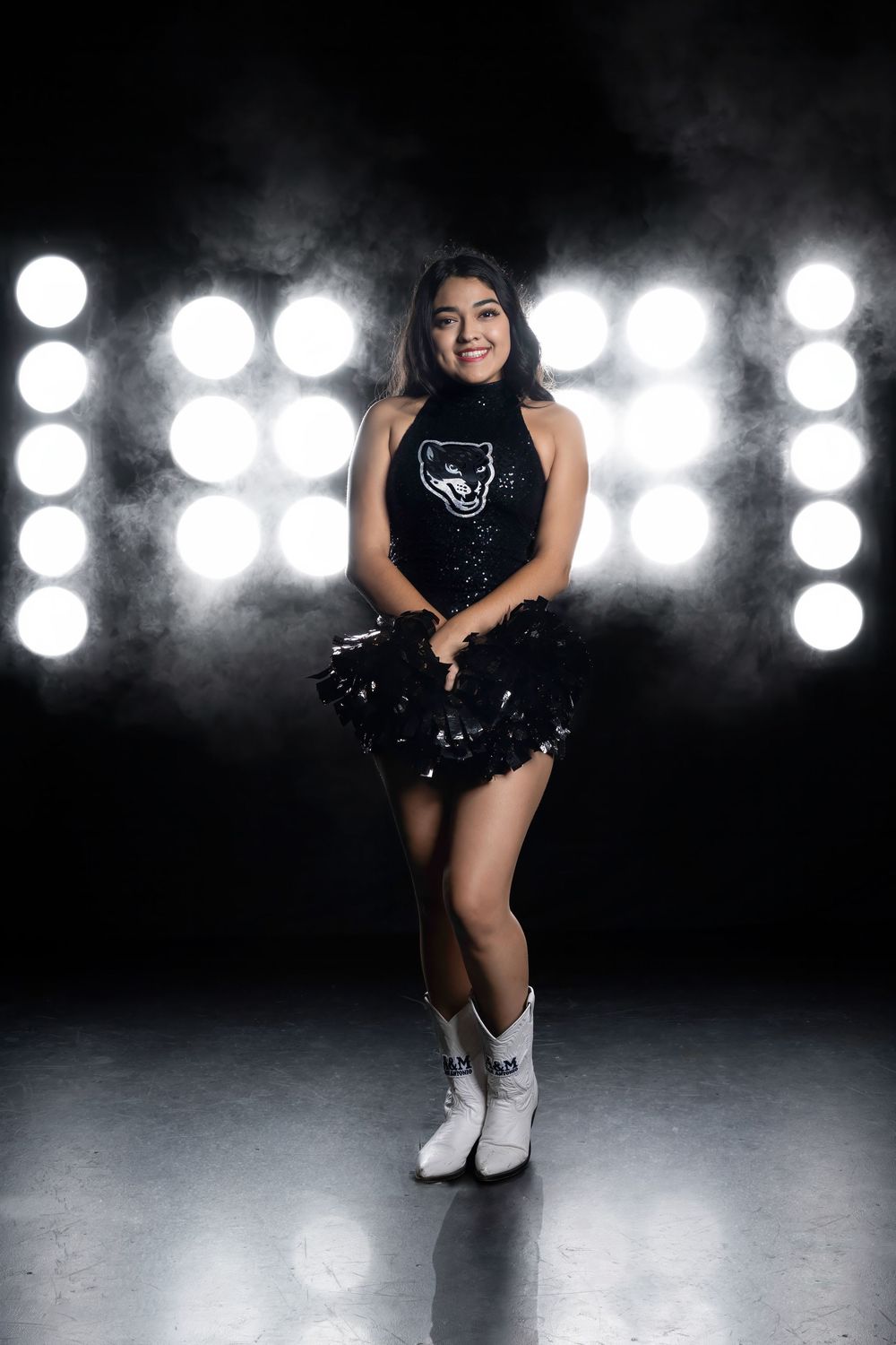 Ice skater poses in sparkly black costume and white boots against dramatic backdrop of bright stage lights.