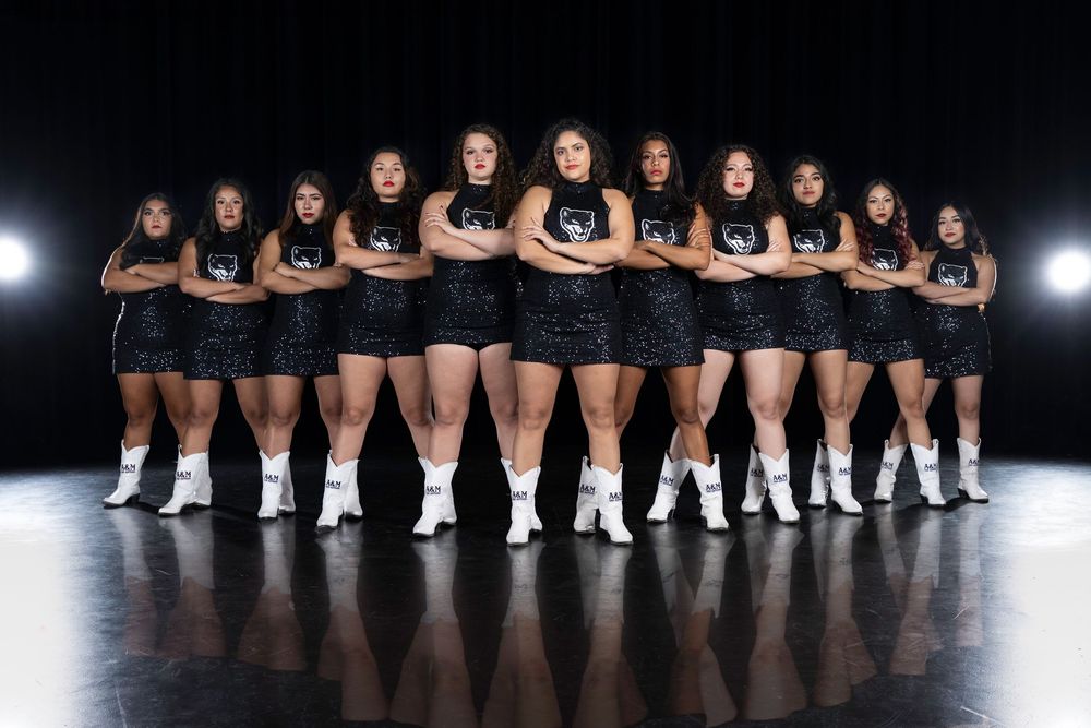 A team of athletes in black uniforms and white shoes pose in formation on a dark stage with dramatic lighting.
