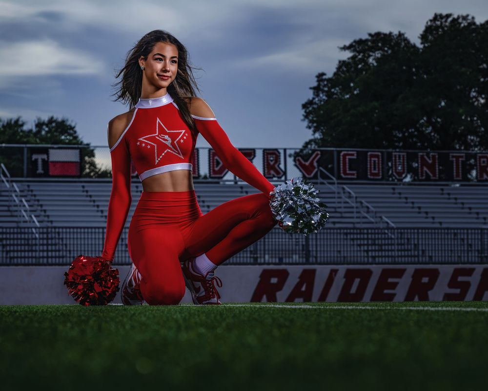 Five cheerleaders in red uniforms pose with pom-poms in front of stadium bleachers during an outdoor photoshoot at dusk.