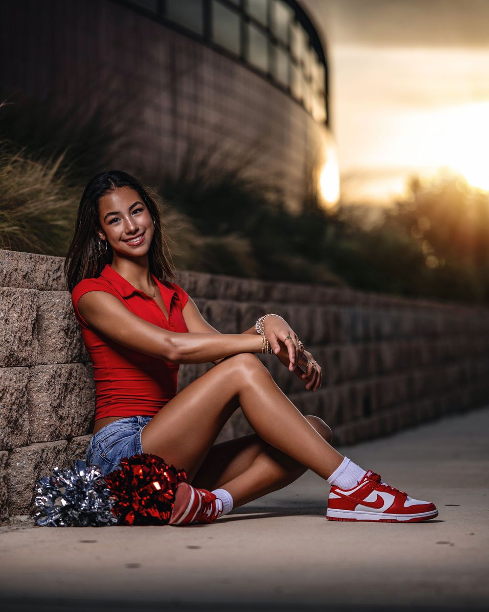 Cheerleader poses in red top and Nike sneakers at sunset against stone wall with pom poms beside her.