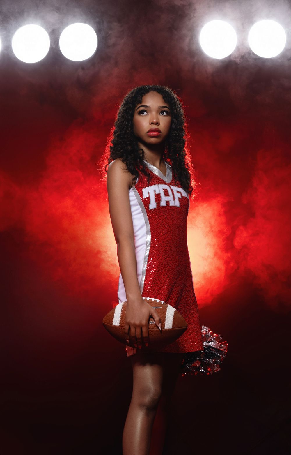 Dramatic portrait of a cheerleader in red and white uniform against a dark red smoky background with bright spotlights.