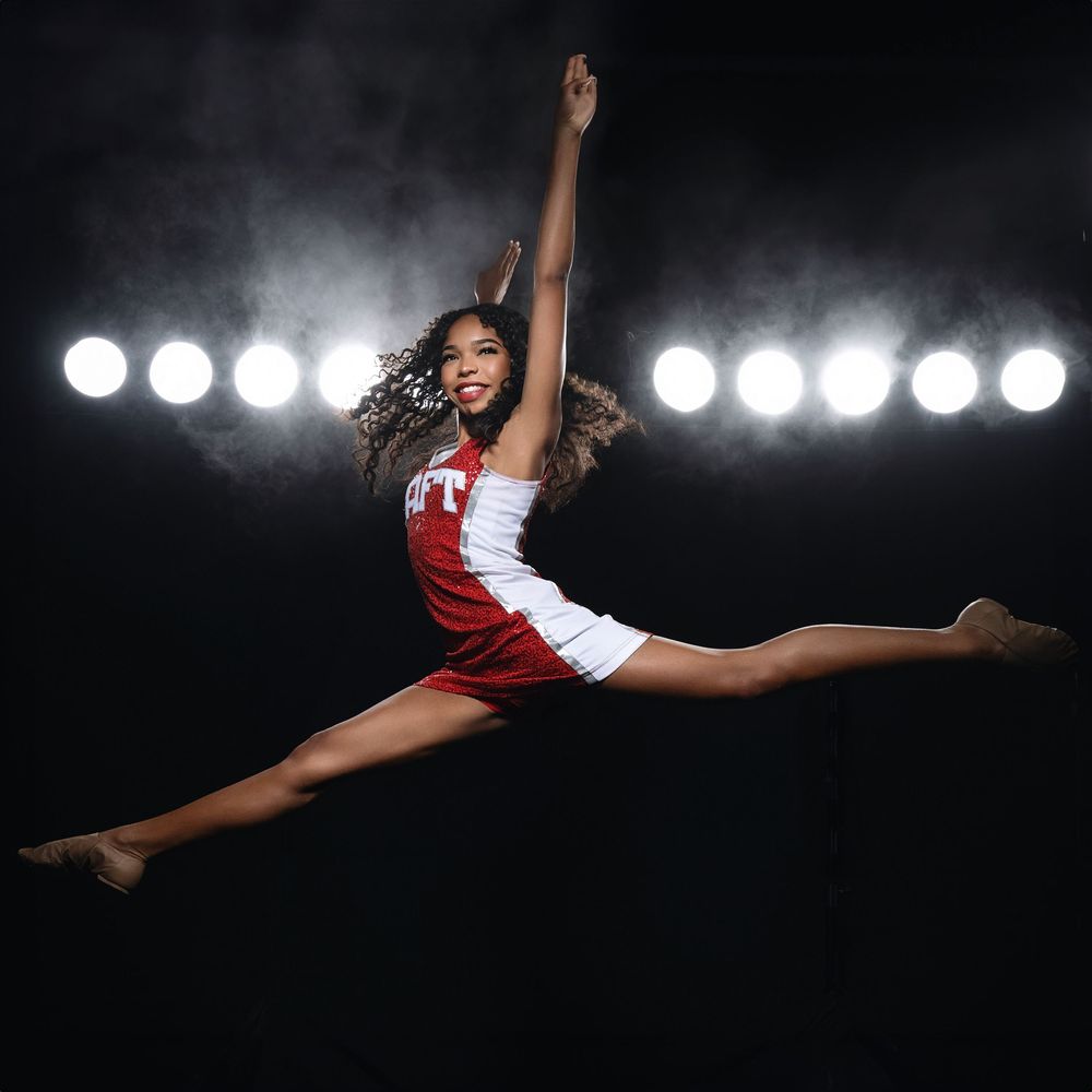 A cheerleader in red and white uniform performs a dramatic split jump in mid-air against spotlights on dark background.