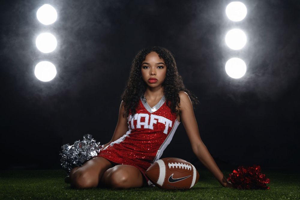 High school cheerleader in red uniform poses on football field at night with pom poms and football.