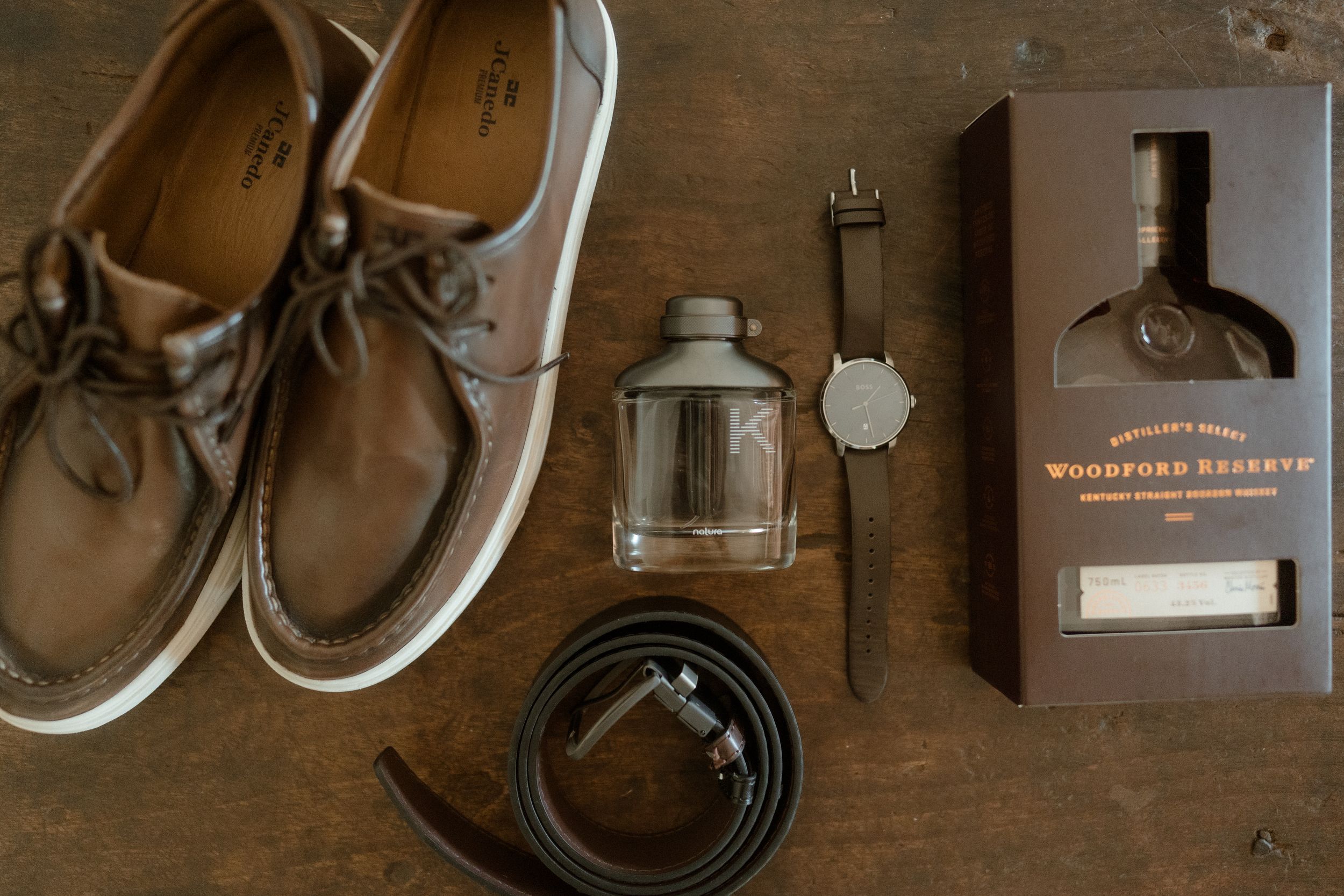 Wedding day accessories including boat shoes, watch, belt and whiskey bottle arranged on wooden surface.