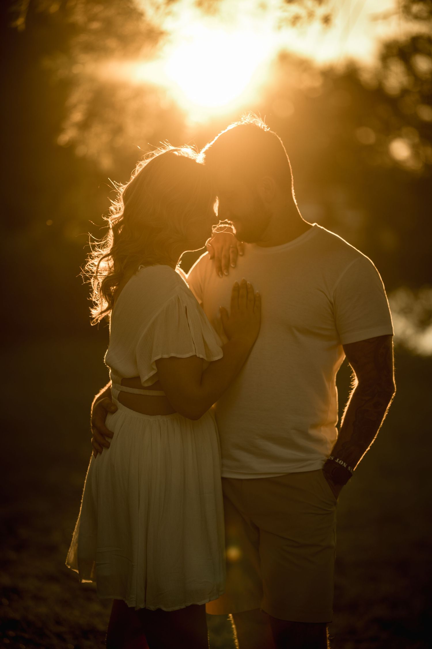 Silhouetted couple embracing in romantic golden sunset light with woman in flowy dress and man in white tshirt.