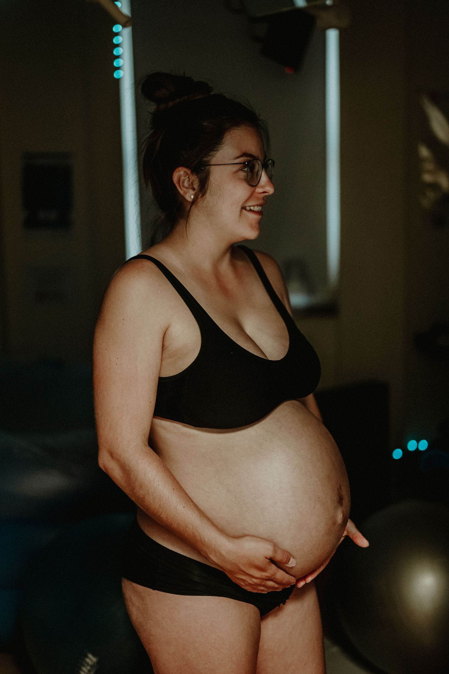 A pregnant person wearing black workout attire stands in a dimly lit room with a gentle smile.