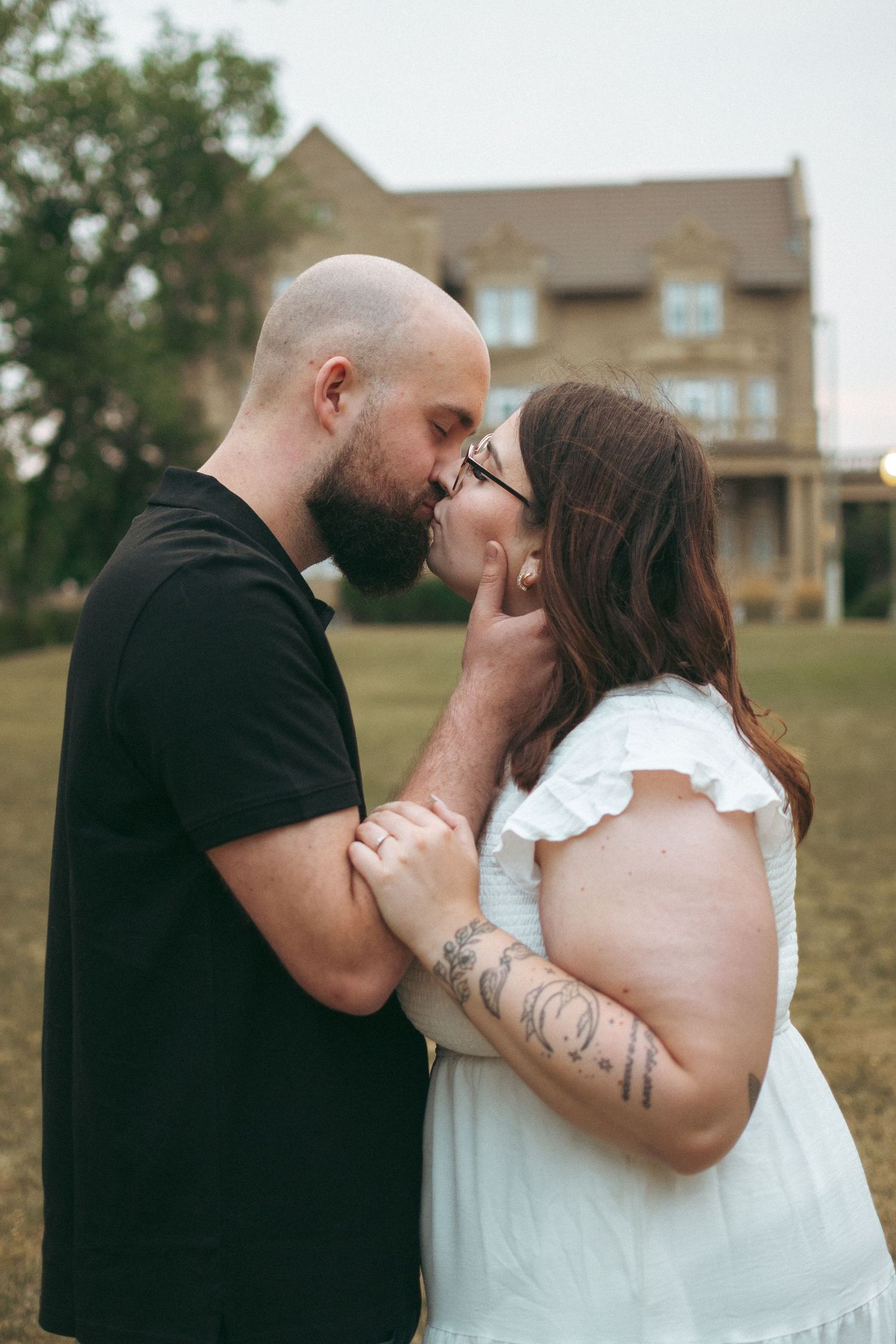 A romantic couple embraces in front of a large house on a grassy lawn during an outdoor photography session.