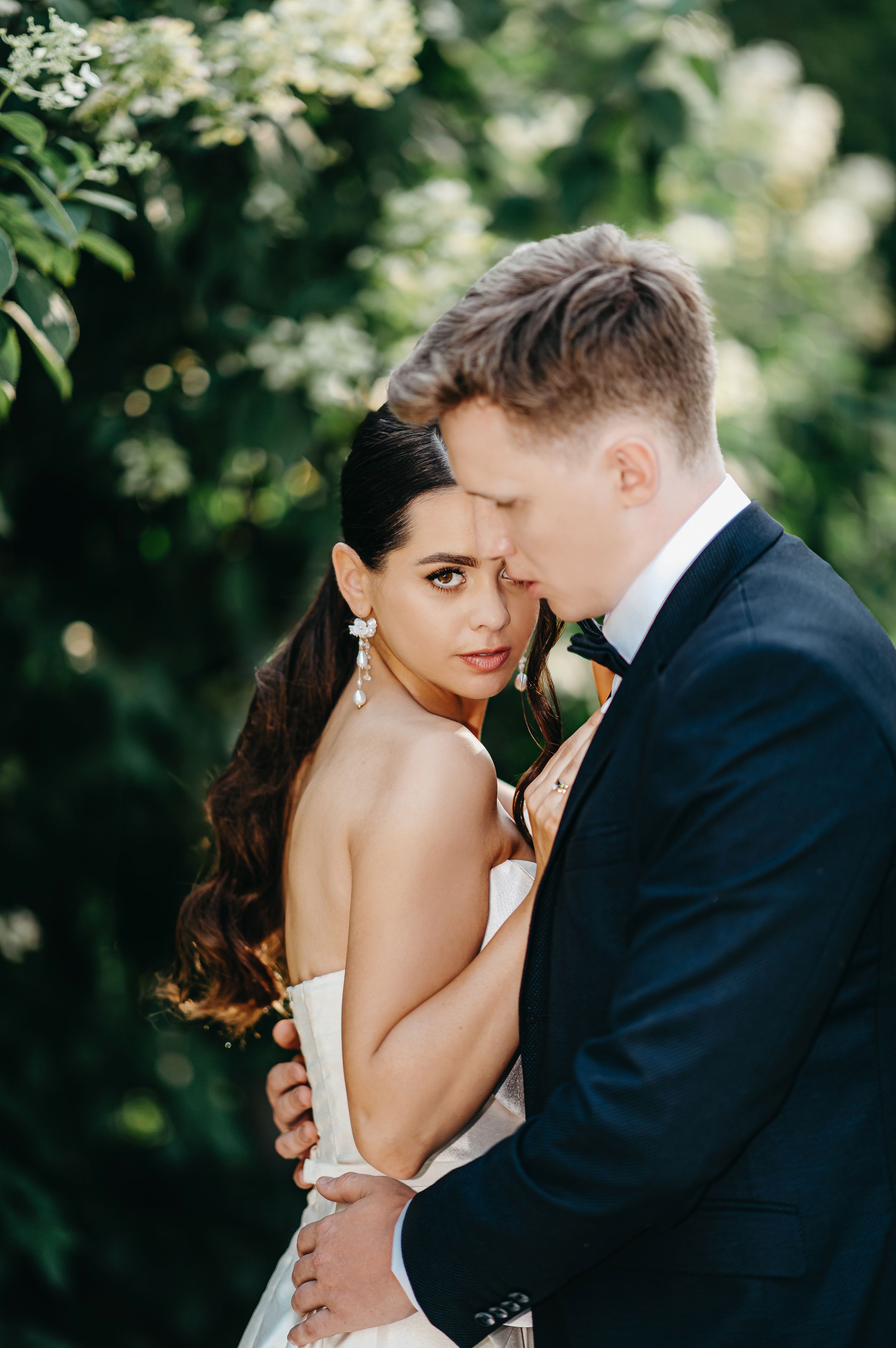 Romantic wedding portrait of couple embracing outdoors with soft bokeh lighting and greenery in background.