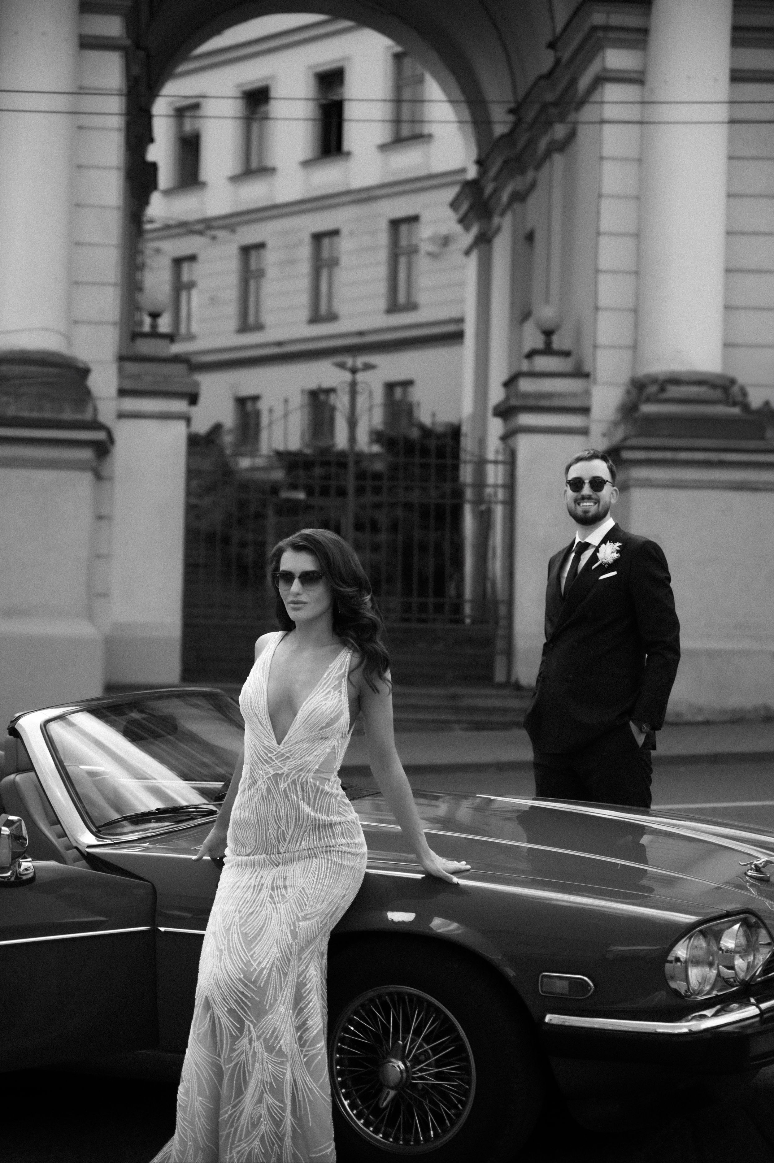 Black and white photo of a couple posing with a classic car in front of an elegant building with columns.