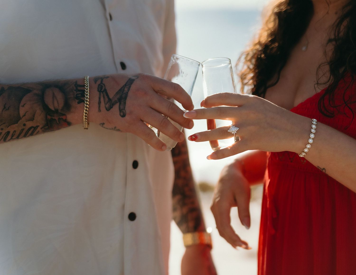 Two people holding champagne glasses together outdoors with one person showing visible arm tattoos.