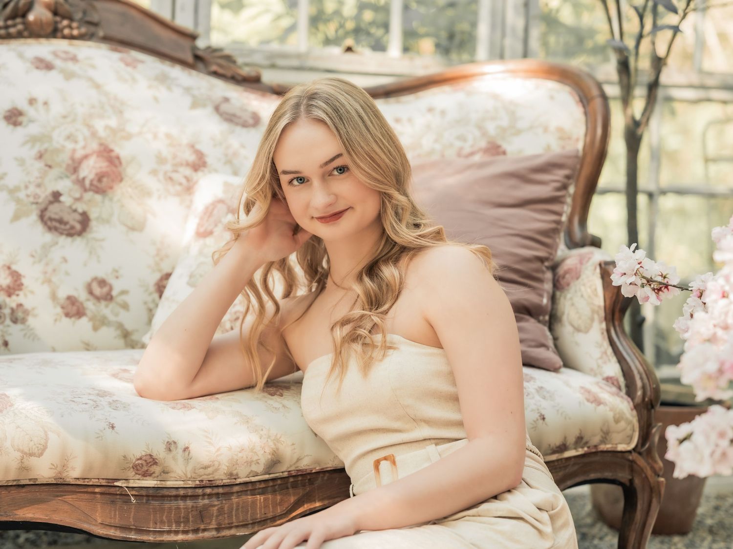 A graceful figure in cream-colored dress reclines on a vintage floral couch in a soft, romantic setting.