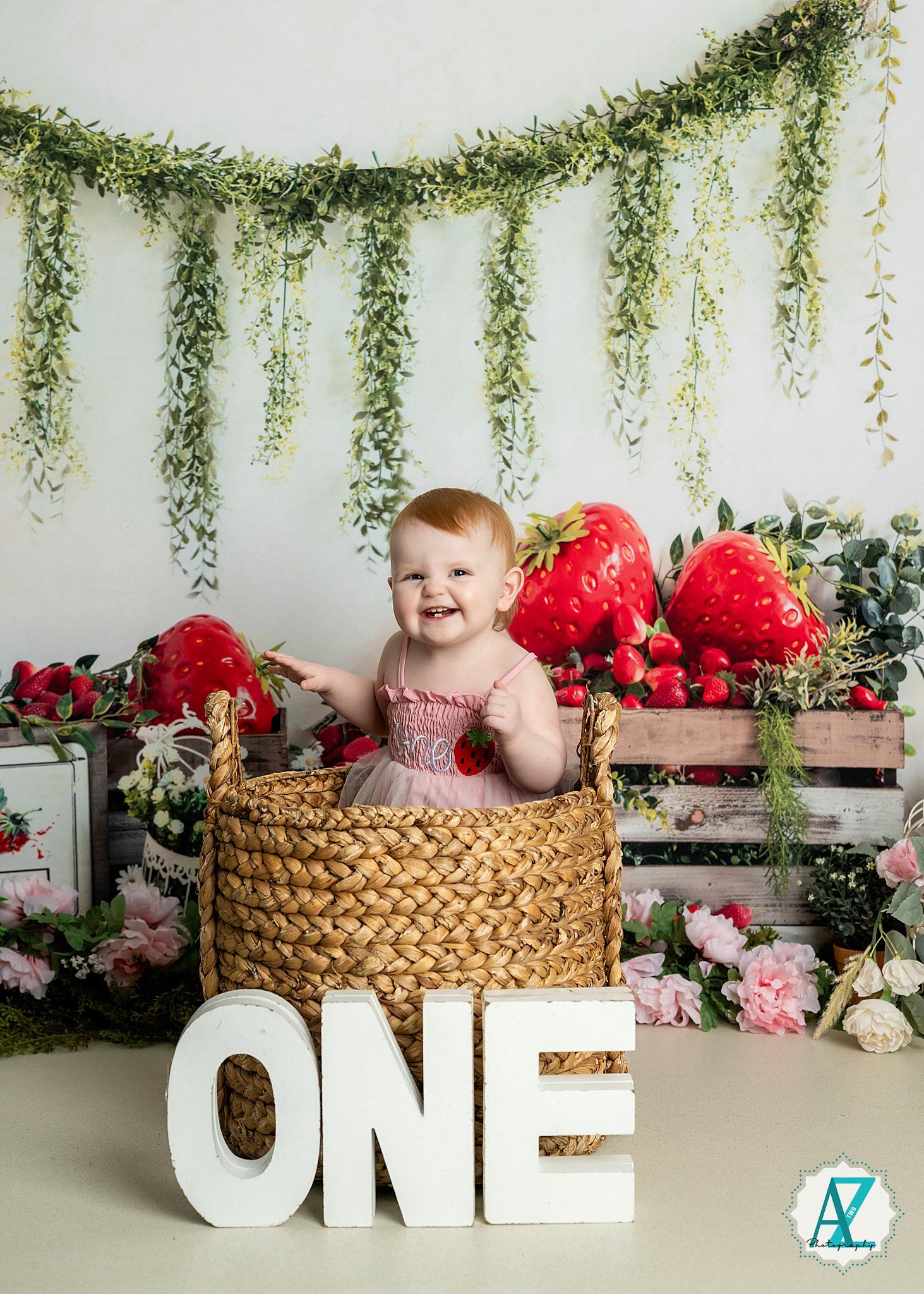 A first birthday photoshoot setup with strawberry decorations, hanging greenery, and a woven basket against a white backdrop.