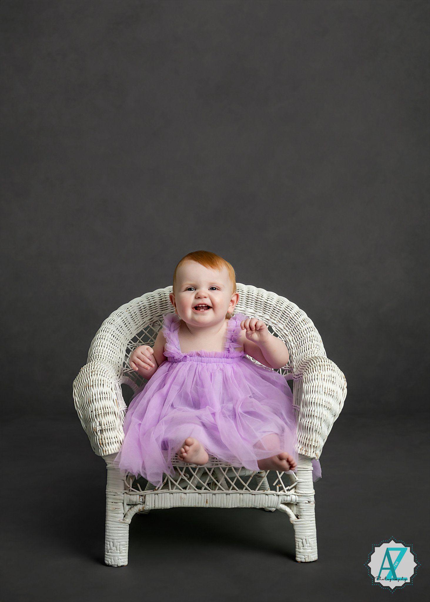 A portrait in a white wicker chair against a dark gray background with purple tulle dress details.