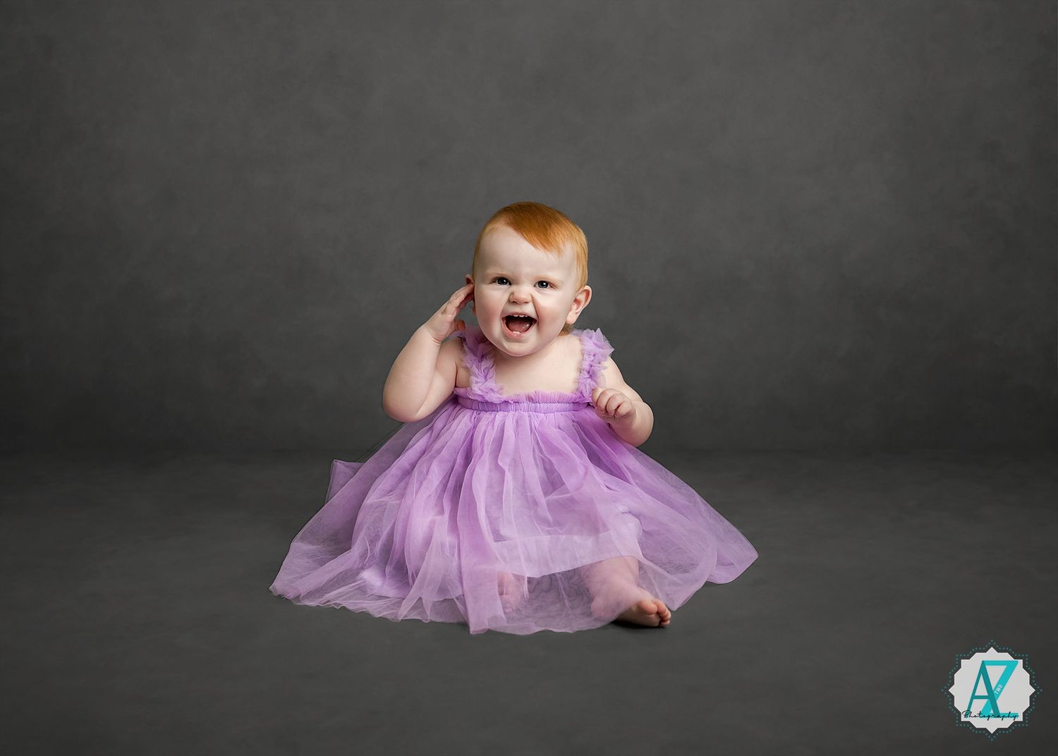 Baby wearing purple tulle dress sits and laughs against dark gray studio background.