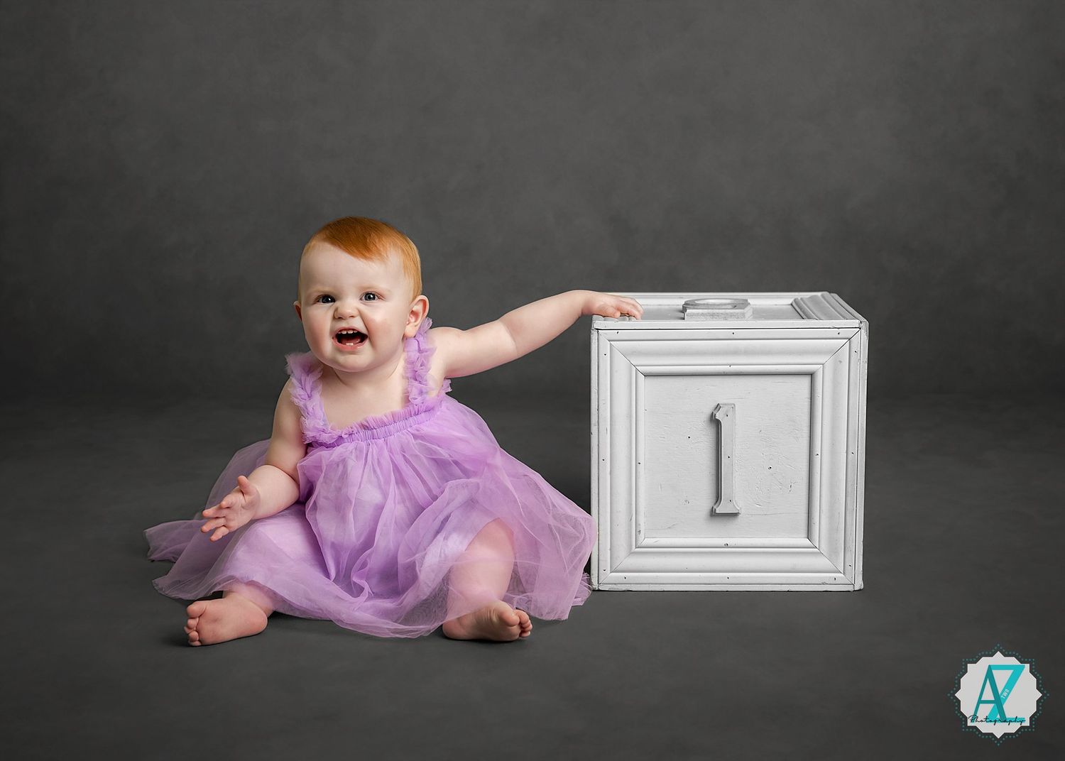 Baby in a purple tulle dress sits on dark floor next to white milestone block during photo session.