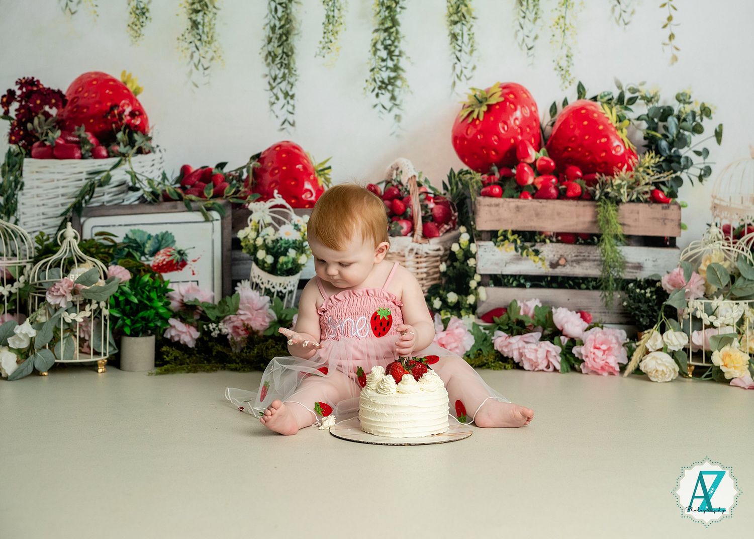 A baby in pink pajamas sits on a white floor with a cake and decorative wooden crates filled with red flowers.