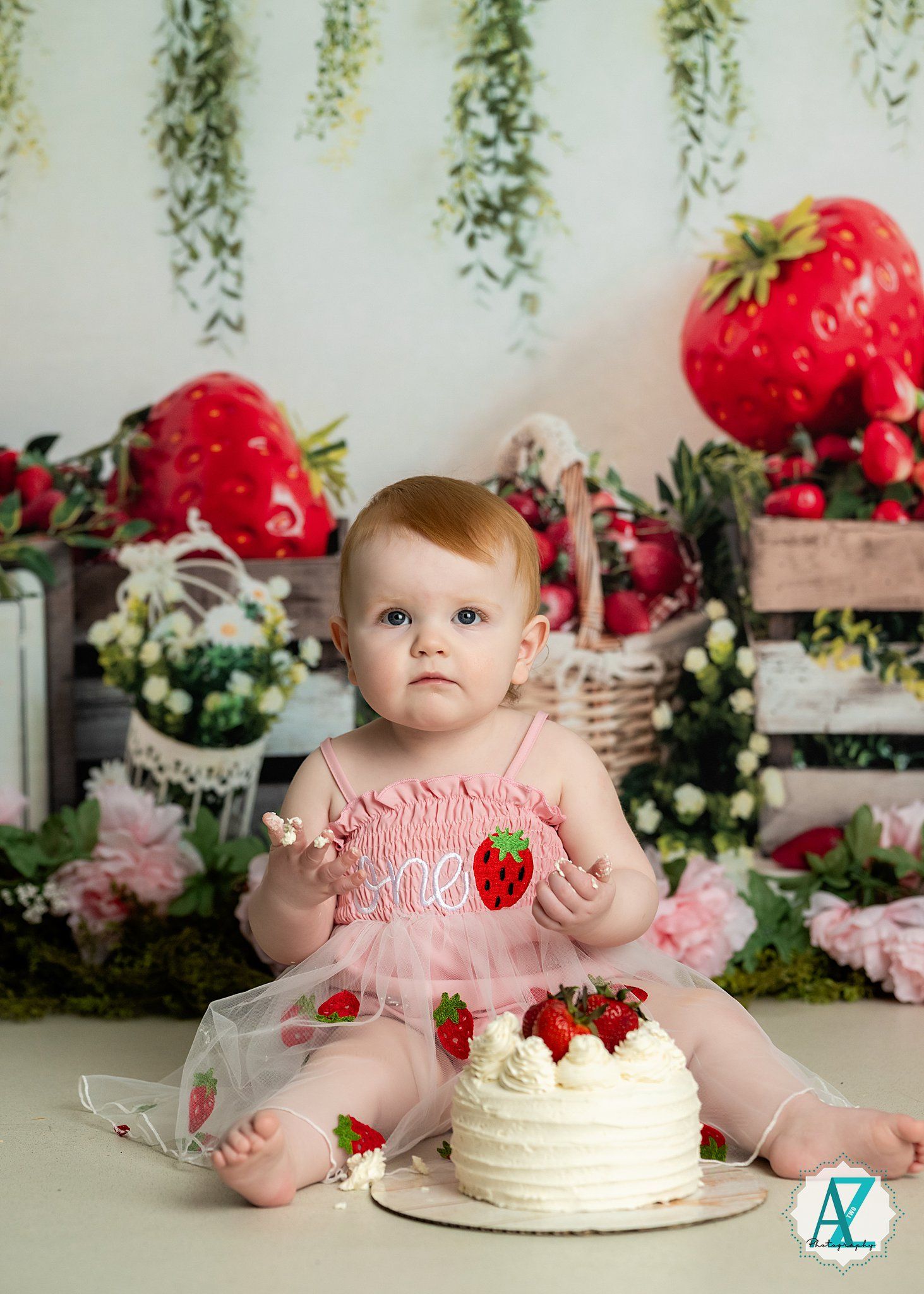 Whimsical cake smash photo setup with giant decorative strawberries, floral arrangements and white frosted cake.