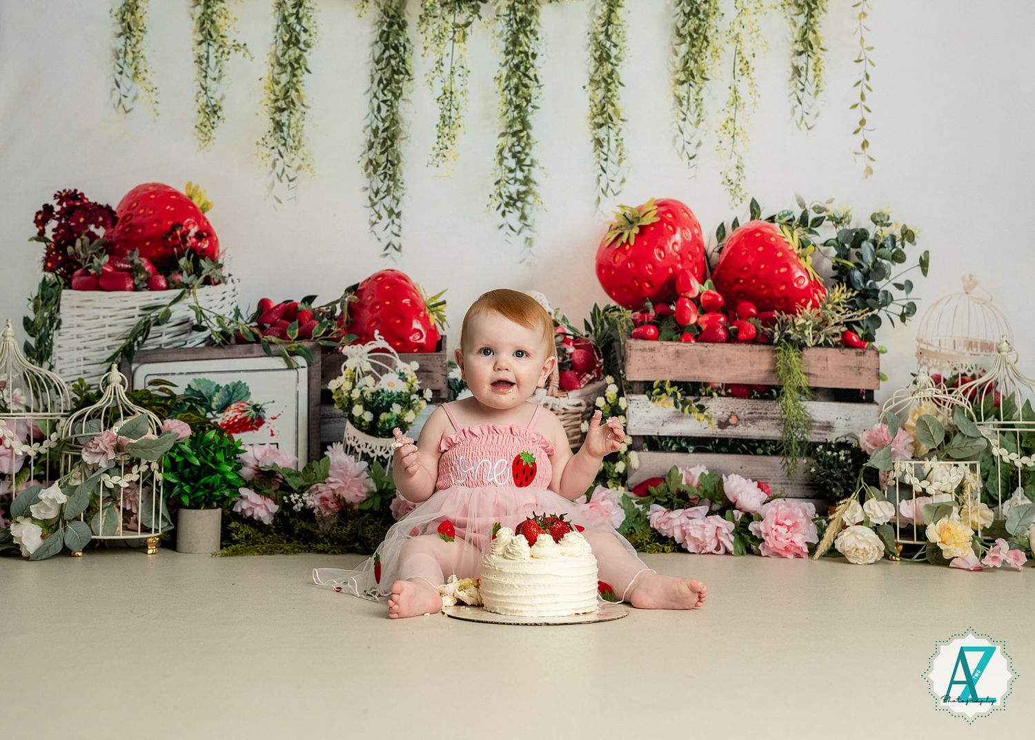 A floral cake smash setup features wooden crates, pink and red flowers, hanging foliage, and a white frosted cake.