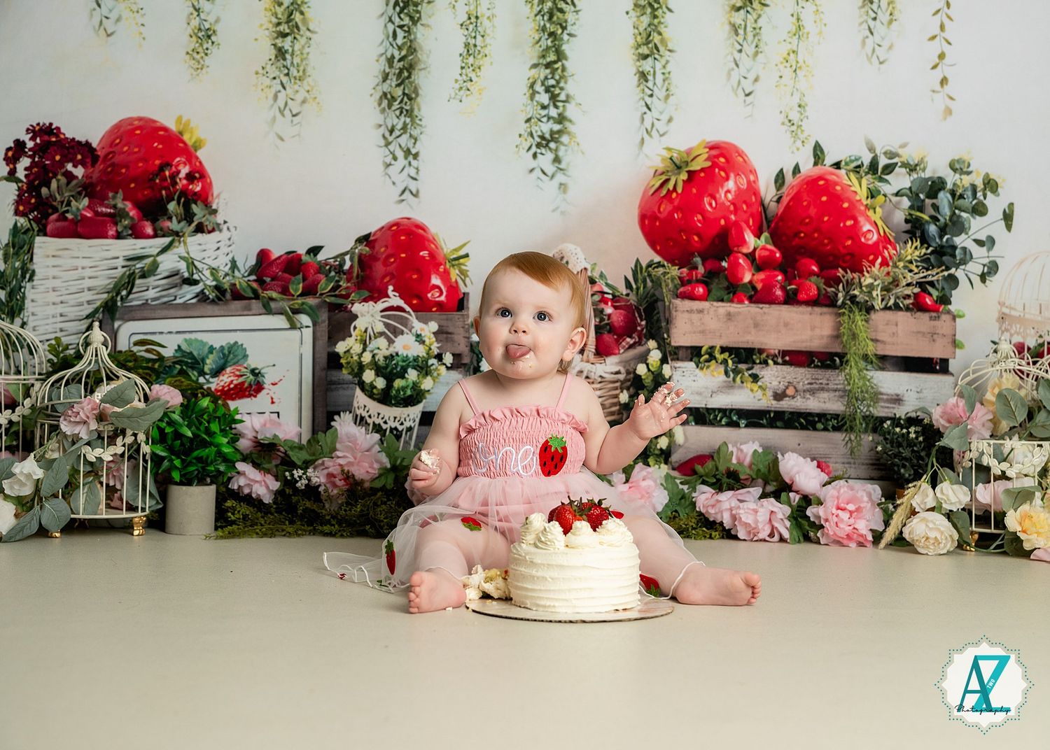 Baby in pink dress with smash cake surrounded by red roses and greenery in vintage style cake smash photo setup.