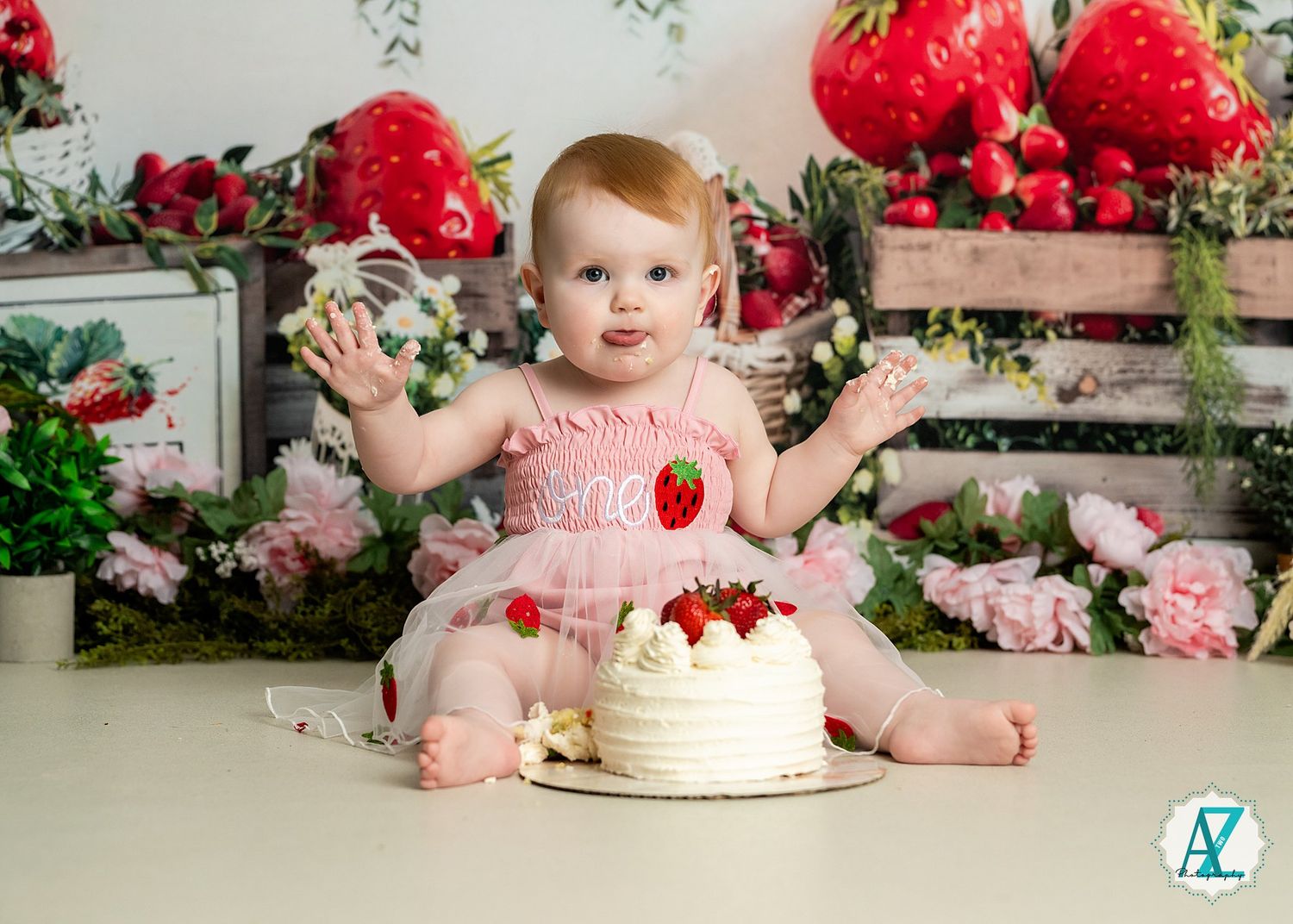 Baby in pink dress sits with white birthday cake surrounded by red and pink floral decorations in studio setting.