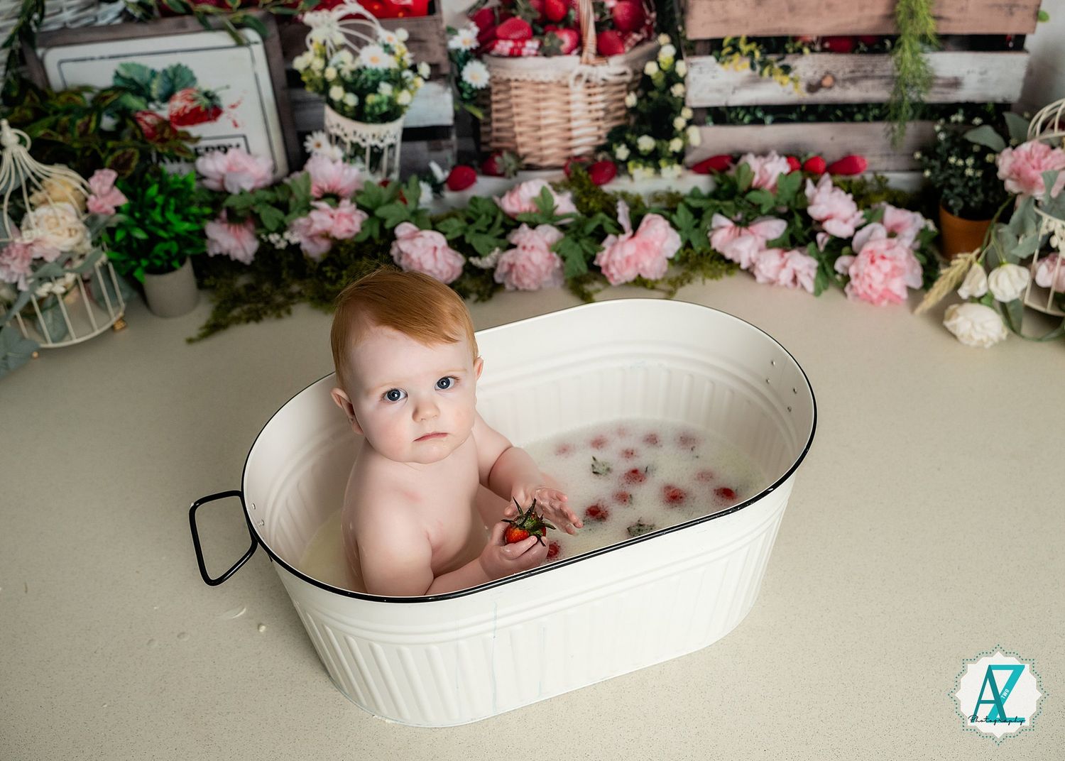 A baby sitting in a vintage white metal tub filled with pink rose petals and surrounded by floral decorations.