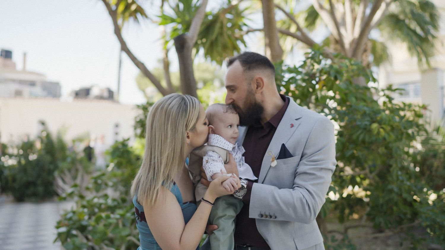 Three people share a tender family moment together outdoors with palm trees in the background.