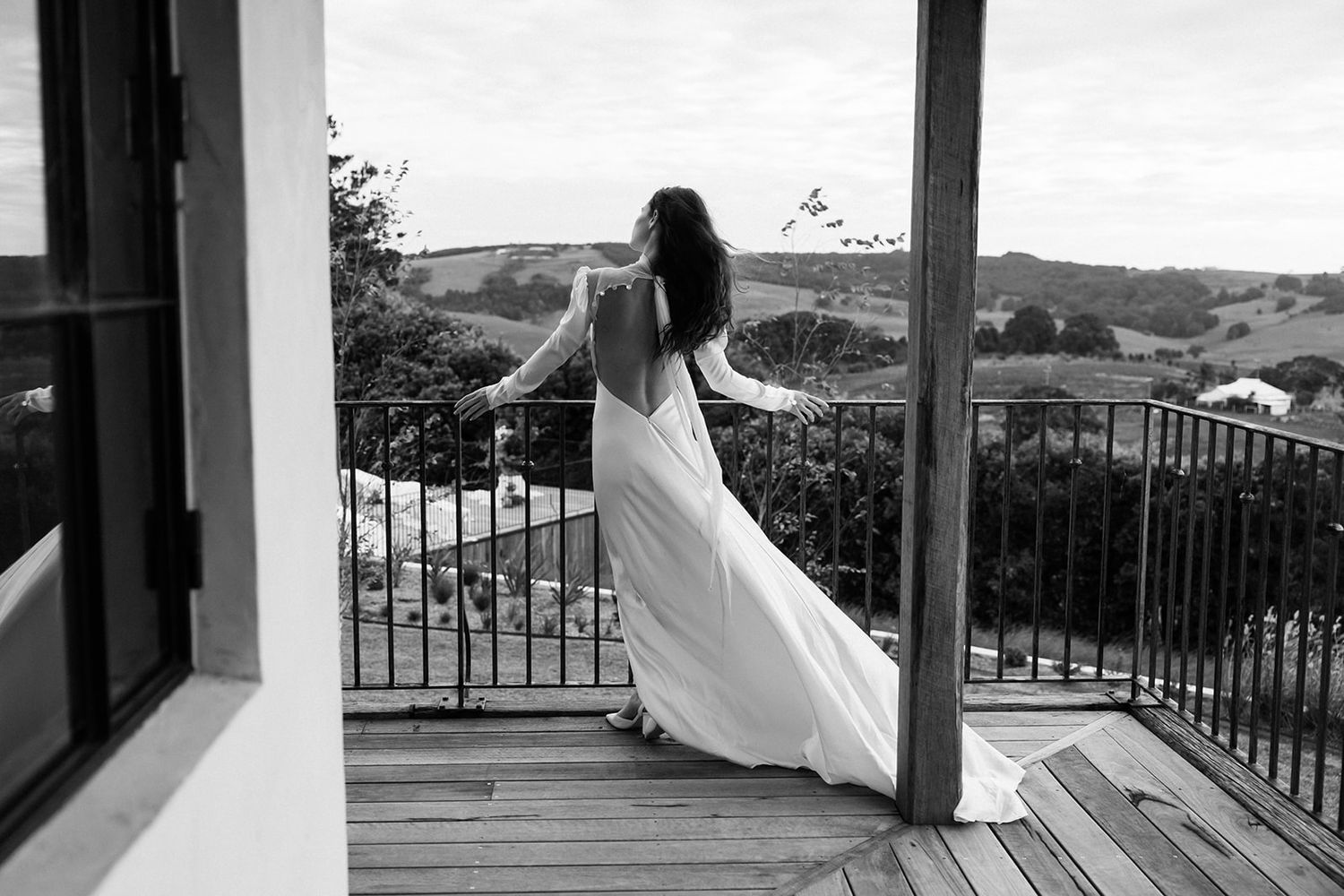 A bride in flowing dress stands on a balcony overlooking rolling hills in dramatic black and white.