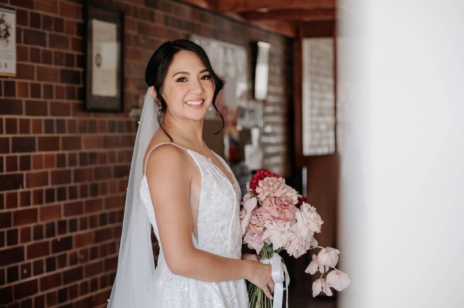 A bride in a white lace wedding dress holds a pink and white bouquet while standing against a brick wall interior.