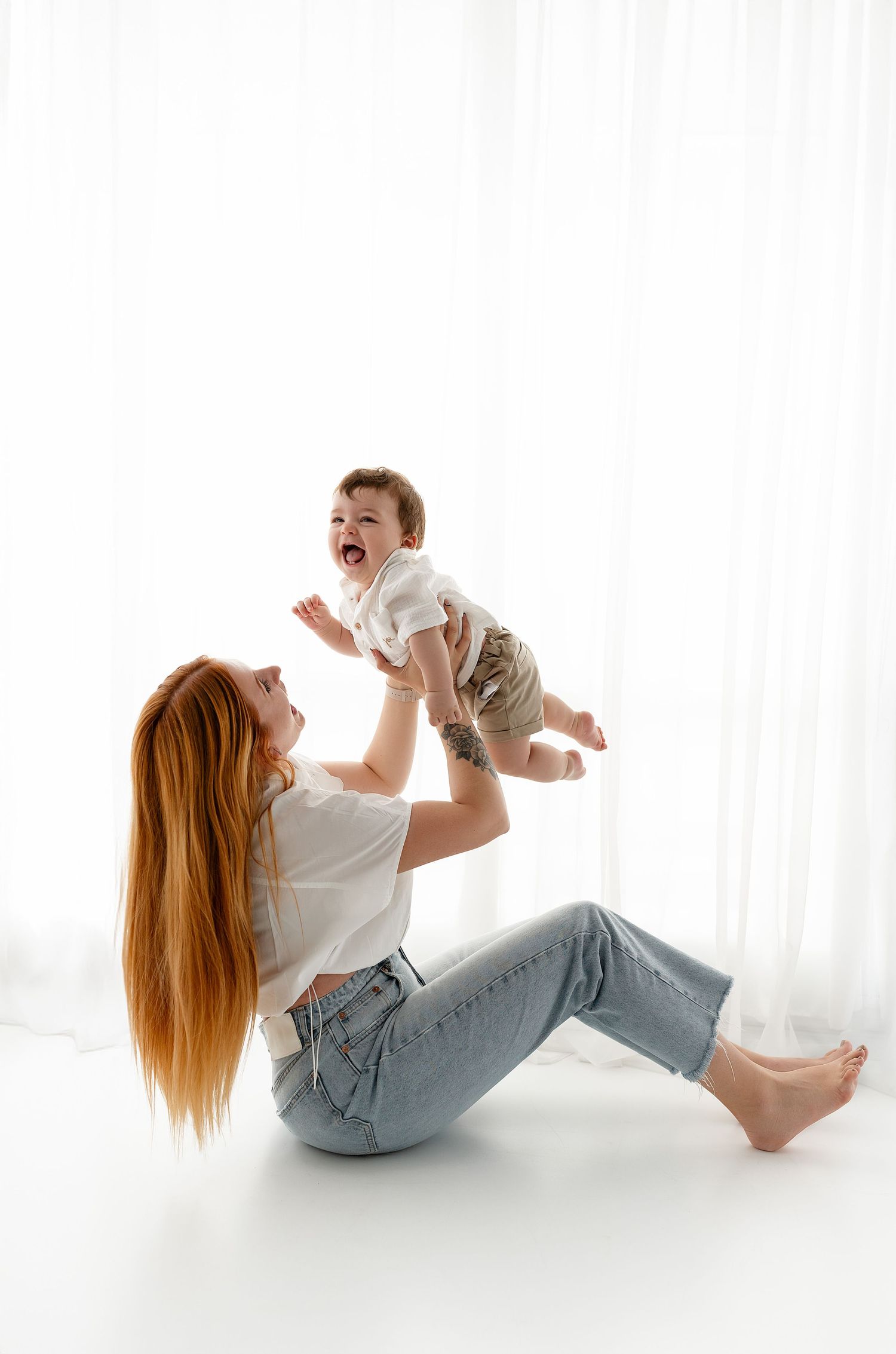 Mother plays with baby on white background, lifting child in air while sitting on floor in casual clothing.
