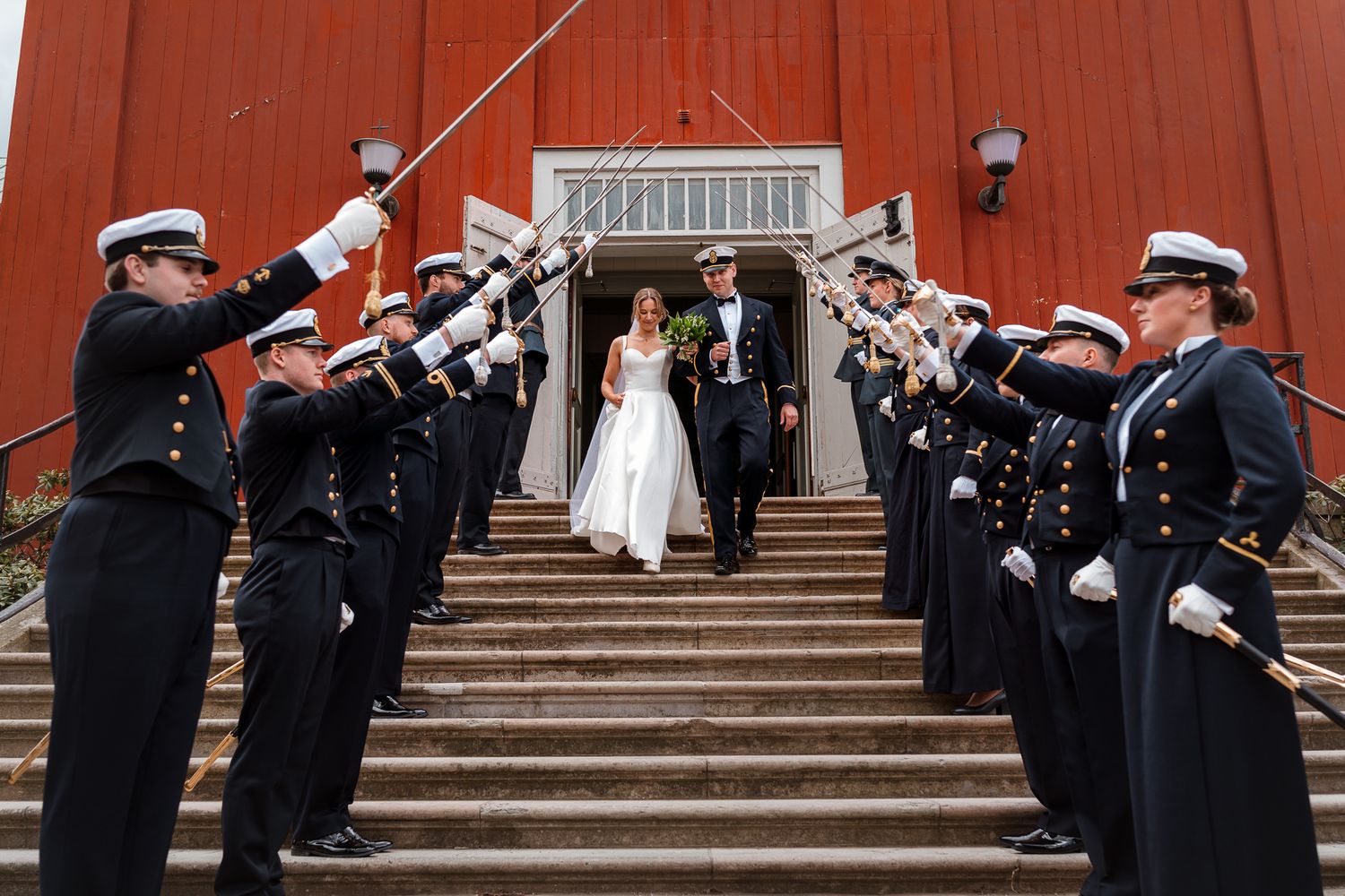 Military honor guard forms saber arch as couple exits red barn building during wedding ceremony.