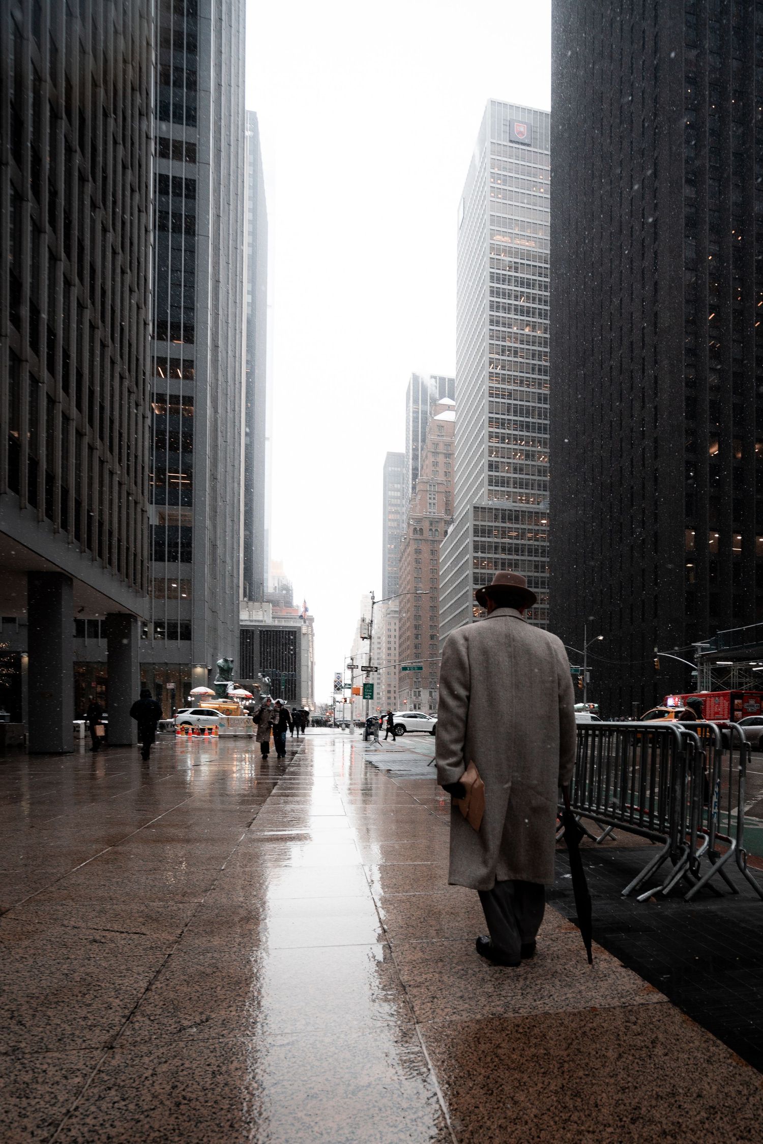A solitary figure in a coat walks on a wet sidewalk between towering skyscrapers in a rainy city.