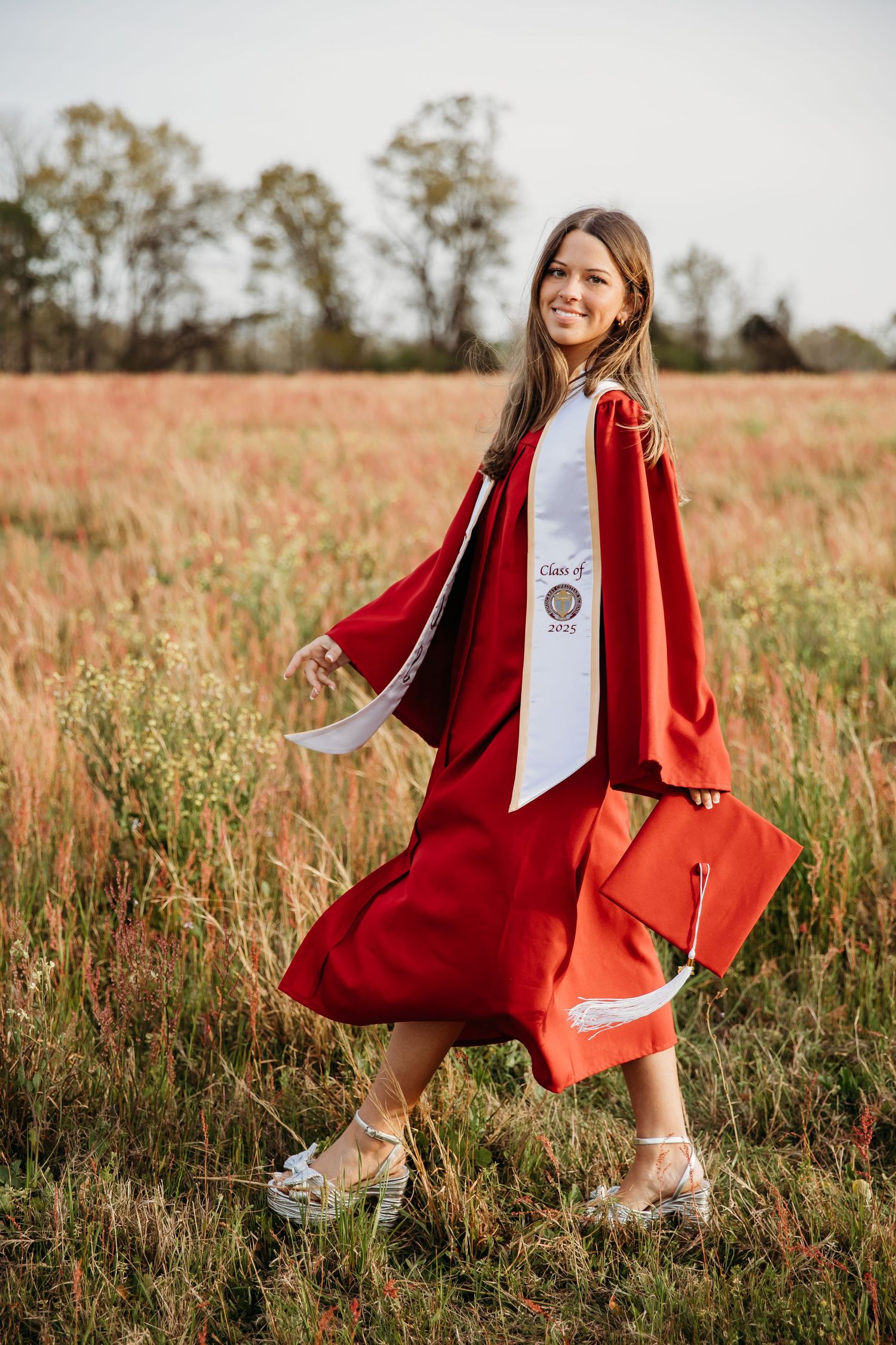 A graduate in red academic regalia poses confidently in a sunny field with an orange tote bag.
