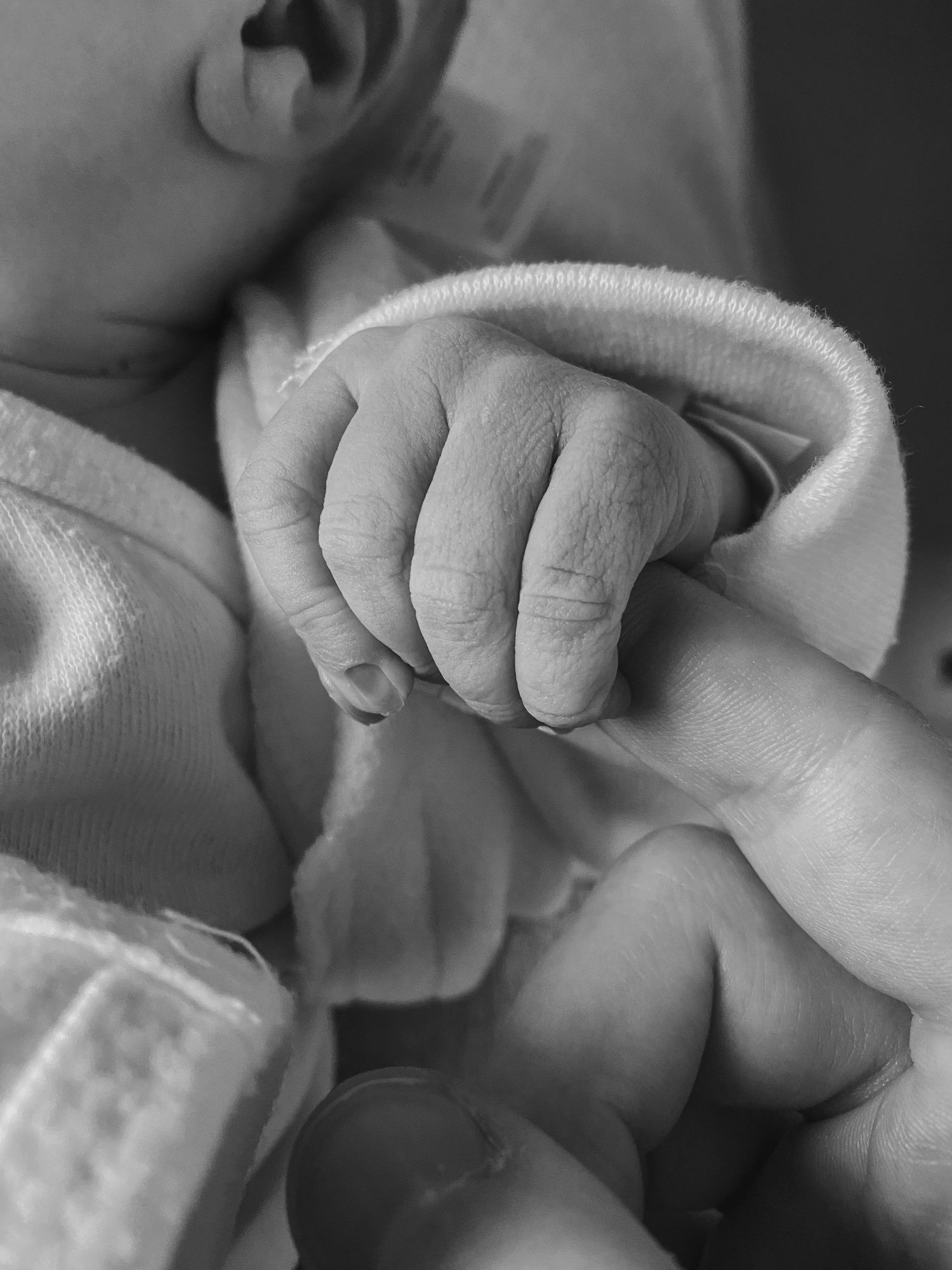 Close-up black and white photo of a tiny baby's hand gripping an adult's finger while wearing white clothing.