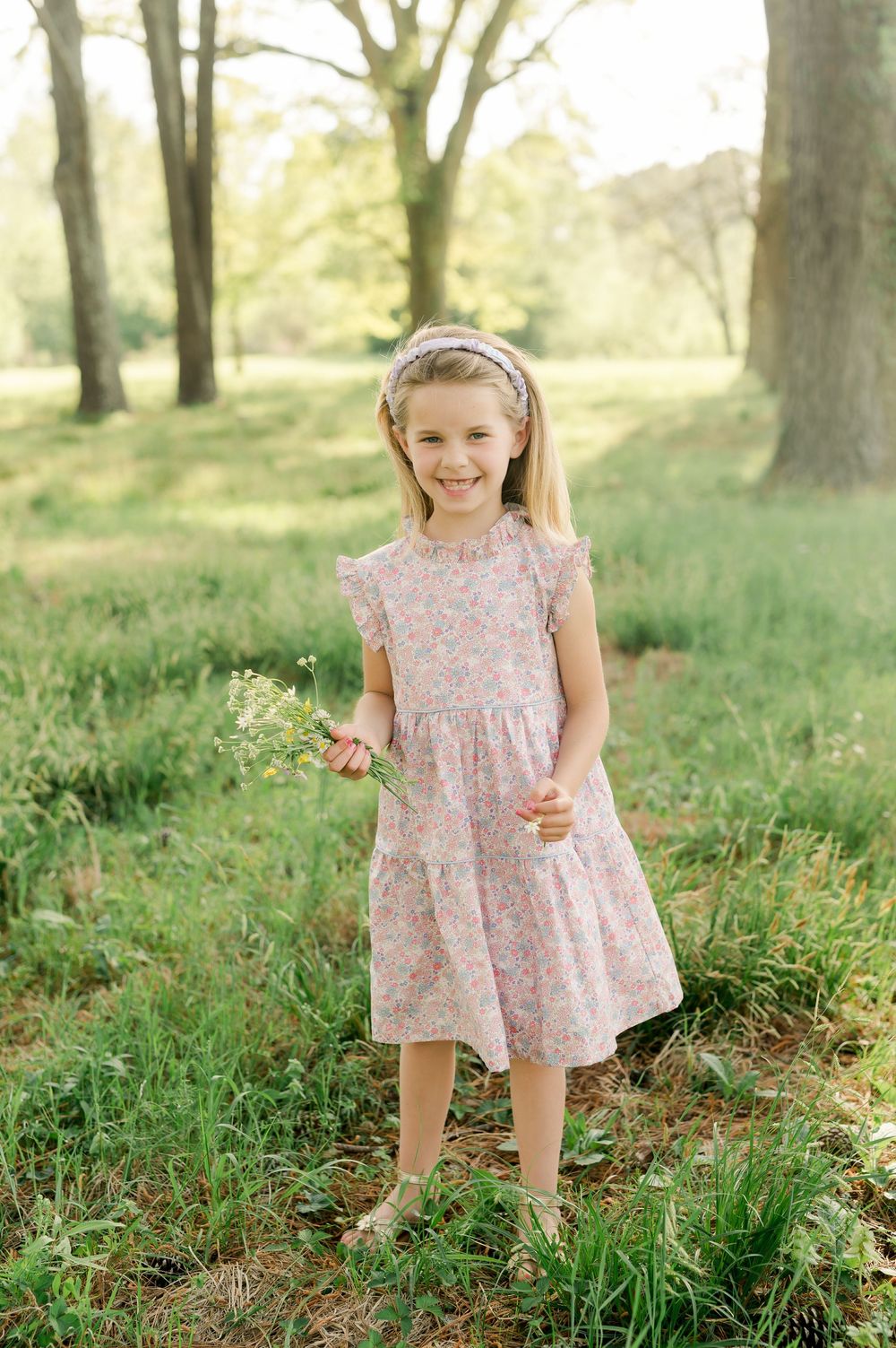 Child in a pink floral dress standing in a sunlit meadow holding wildflowers among tall trees.