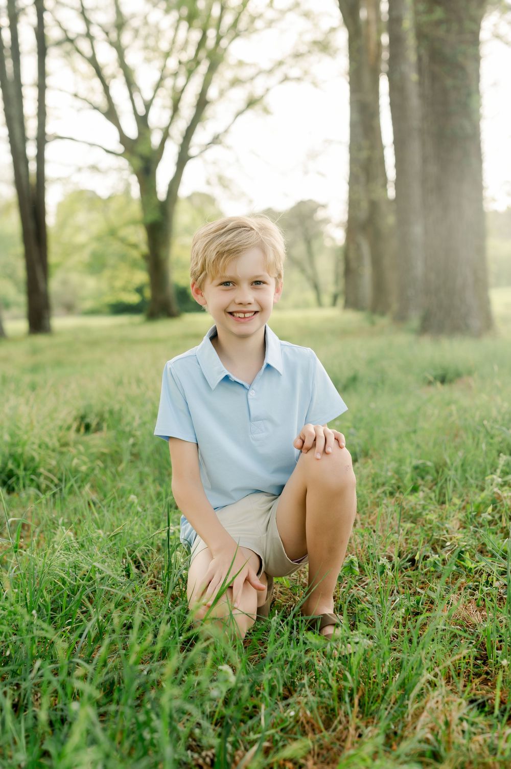 Young person kneeling on grass in a light blue polo shirt and khaki shorts in a sunlit wooded park setting.