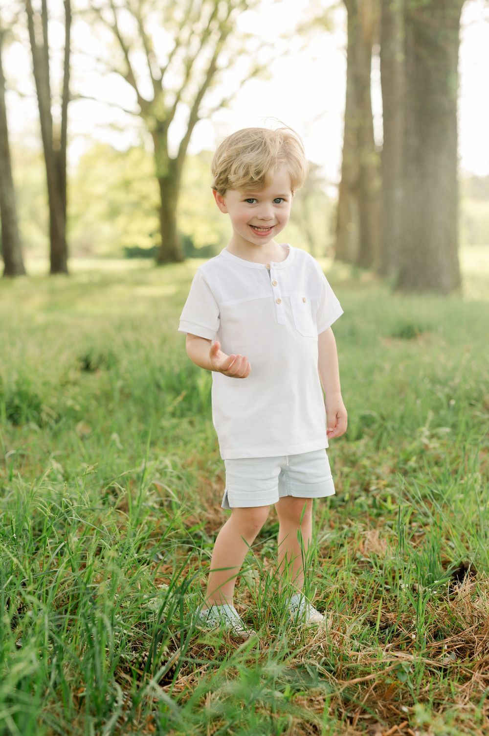 A child in a white polo shirt and light blue shorts stands smiling in a sunlit wooded area with green grass.