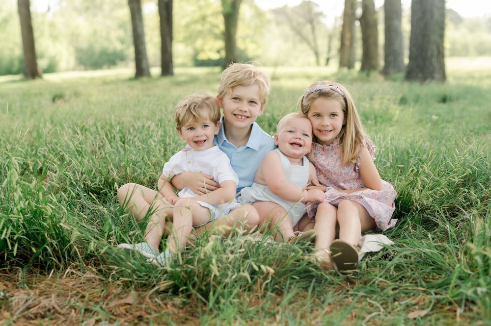 Four young children sitting together in tall grass under a row of trees on a sunny day.