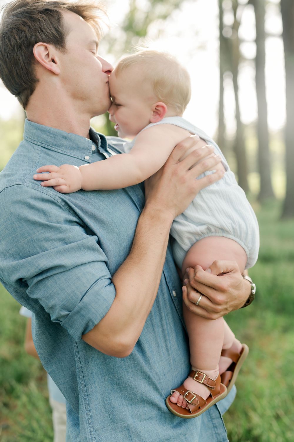 A tender moment between a parent and baby in a denim shirt against a forest backdrop.