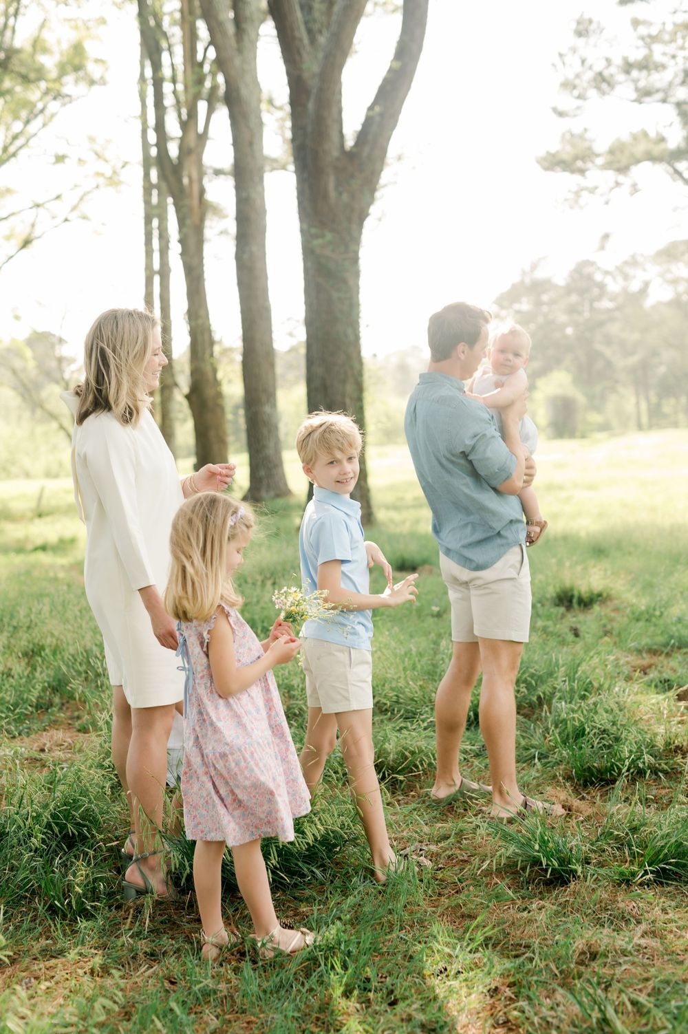 A family stands together outdoors in a sun-dappled wooded area wearing light summer clothing.