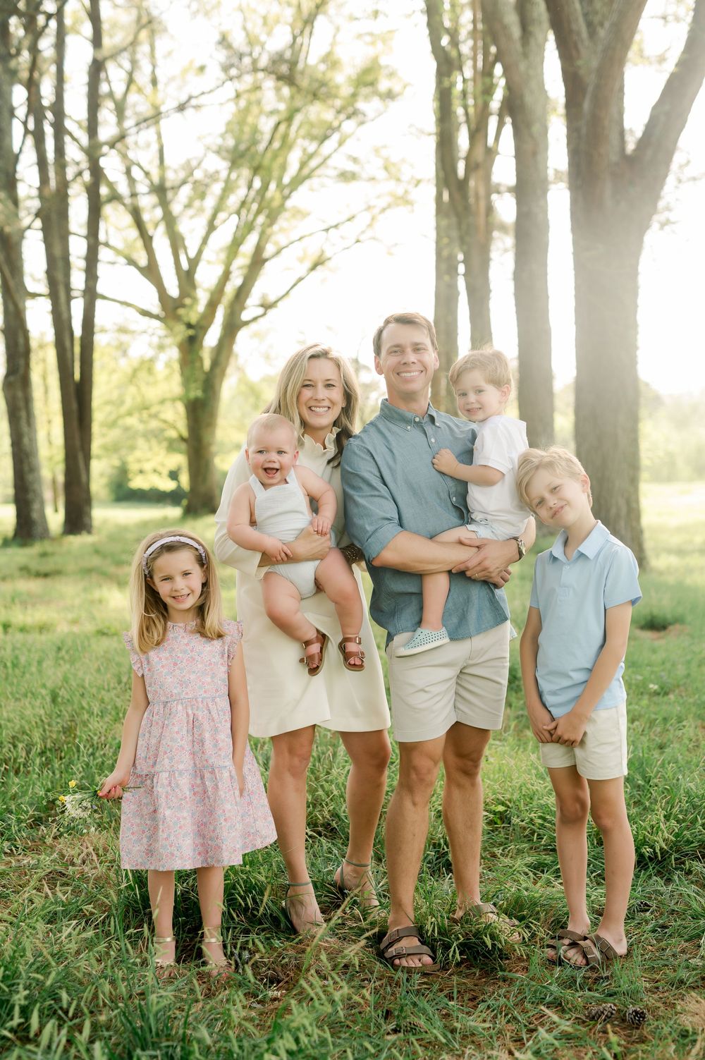 Family of five stands together in front of sunlit trees wearing coordinated pastel spring outfits.