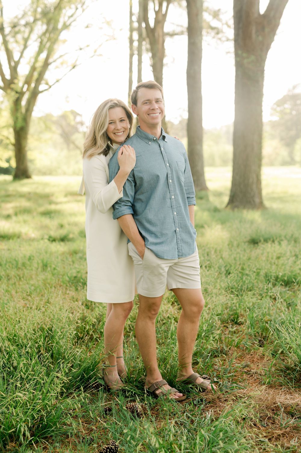 A couple stands together in a sunlit grassy field with tall trees behind them on a warm afternoon.